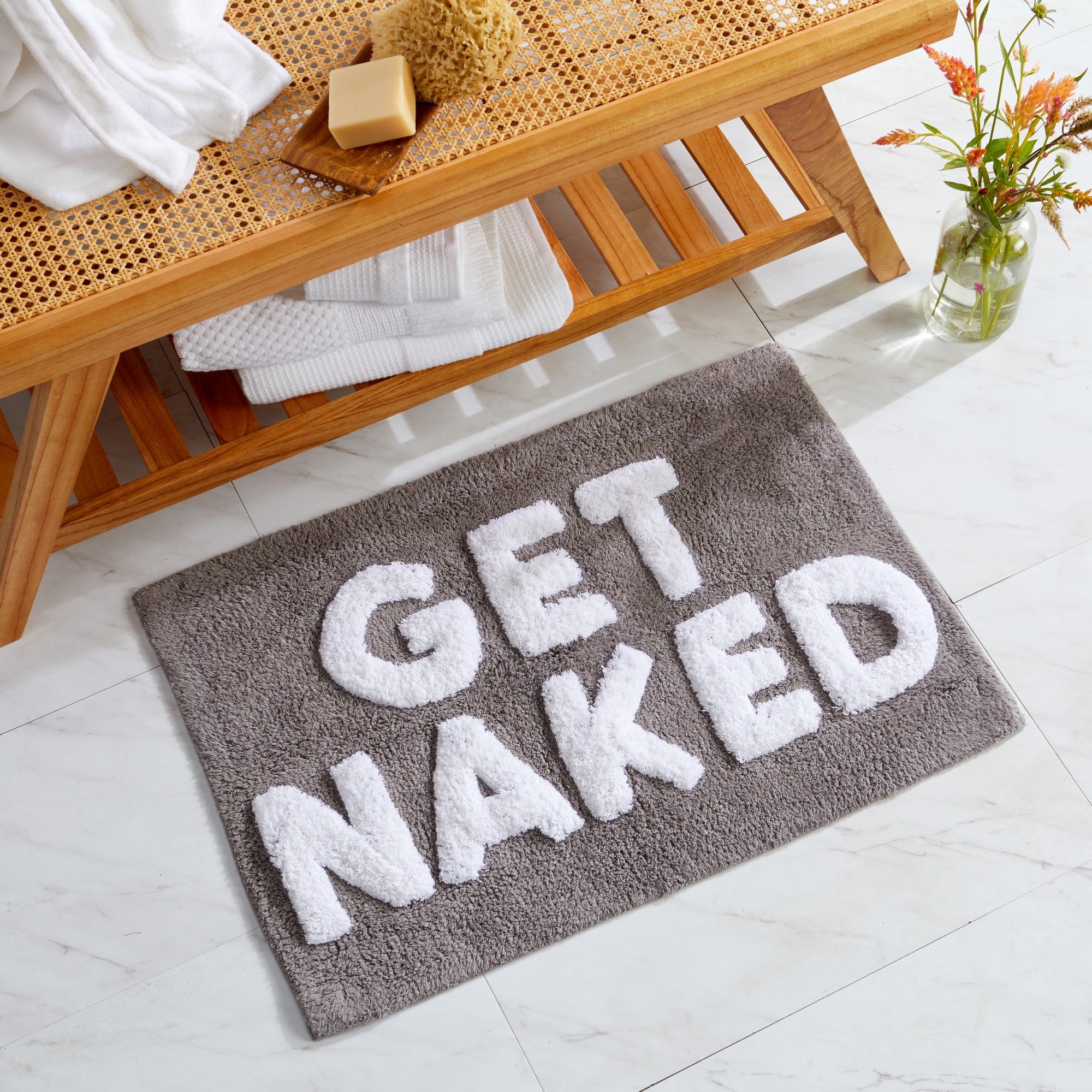 Gray tufted cotton bath rug featuring raised white "GET NAKED" lettering. Soft, plush, and highly absorbent, shown in a modern bathroom setting with white tile flooring and wooden accents. Measures 20 x 30 inches.