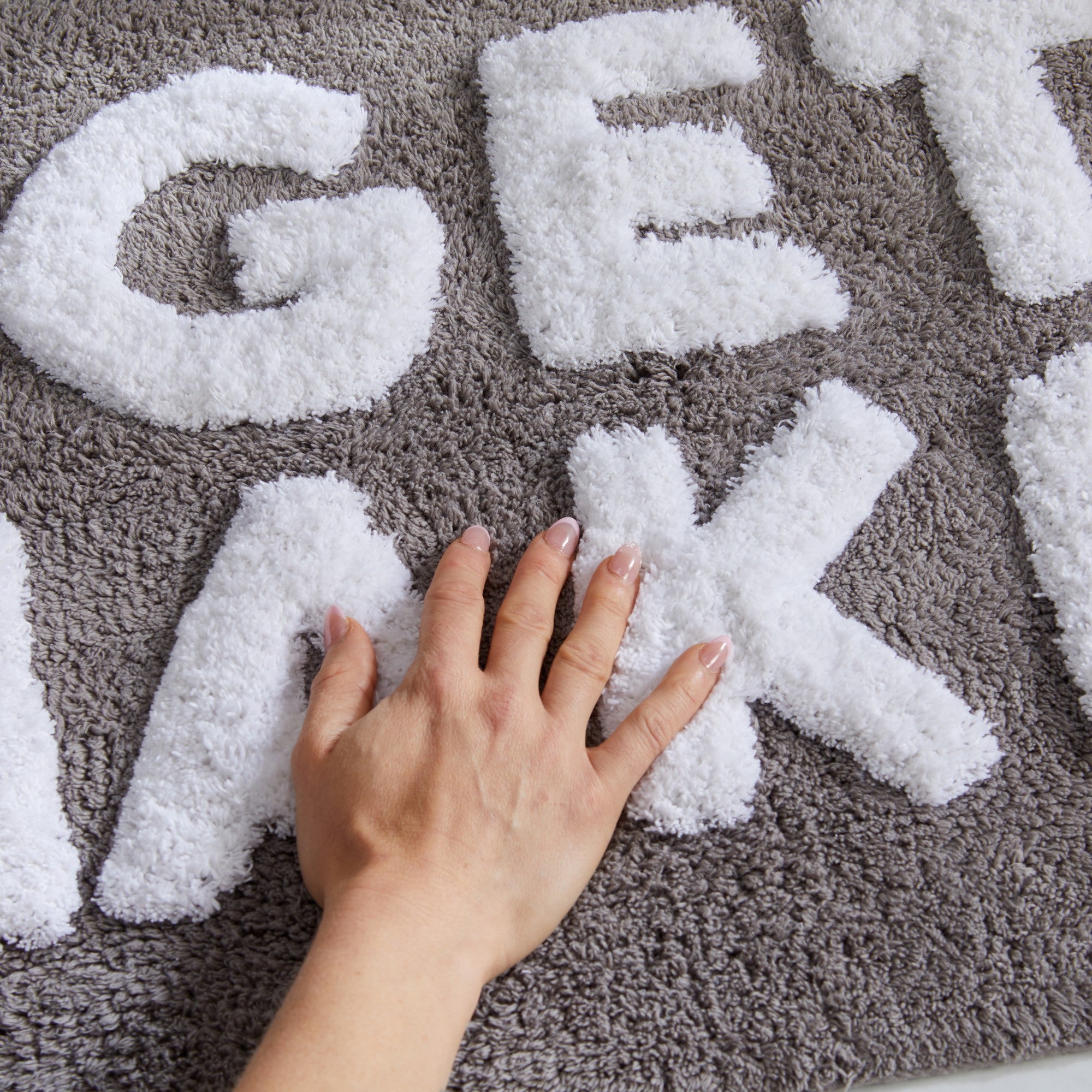 Close-up of a gray tufted cotton bath rug with raised white “GET NAKED” lettering. A hand presses into the plush, soft texture to showcase the rug’s comfort and absorbency.