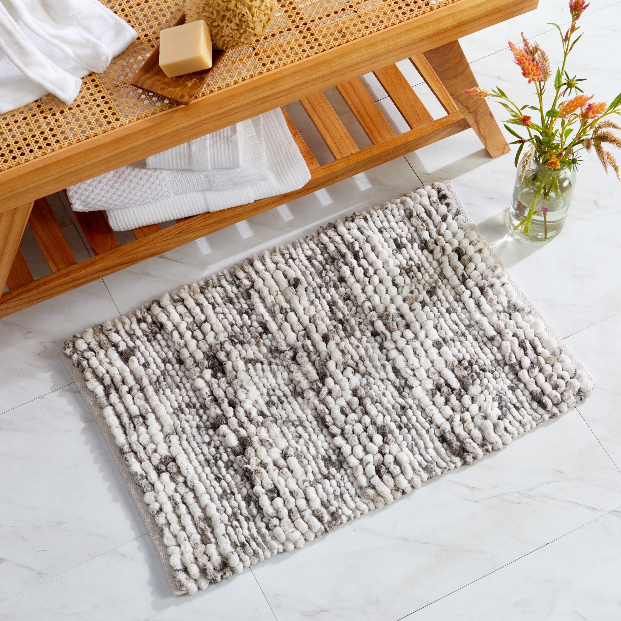 Space-dyed bath rug in neutral ivory and gray tones placed on a bathroom floor beside a natural wood bench. The textured microfiber surface provides a soft, plush feel underfoot while adding warmth, comfort, and modern style to the bathroom décor.