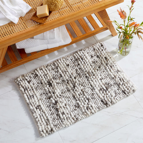 Space-dyed bath rug in neutral ivory and gray tones placed on a bathroom floor beside a natural wood bench. The textured microfiber surface provides a soft, plush feel underfoot while adding warmth, comfort, and modern style to the bathroom décor.