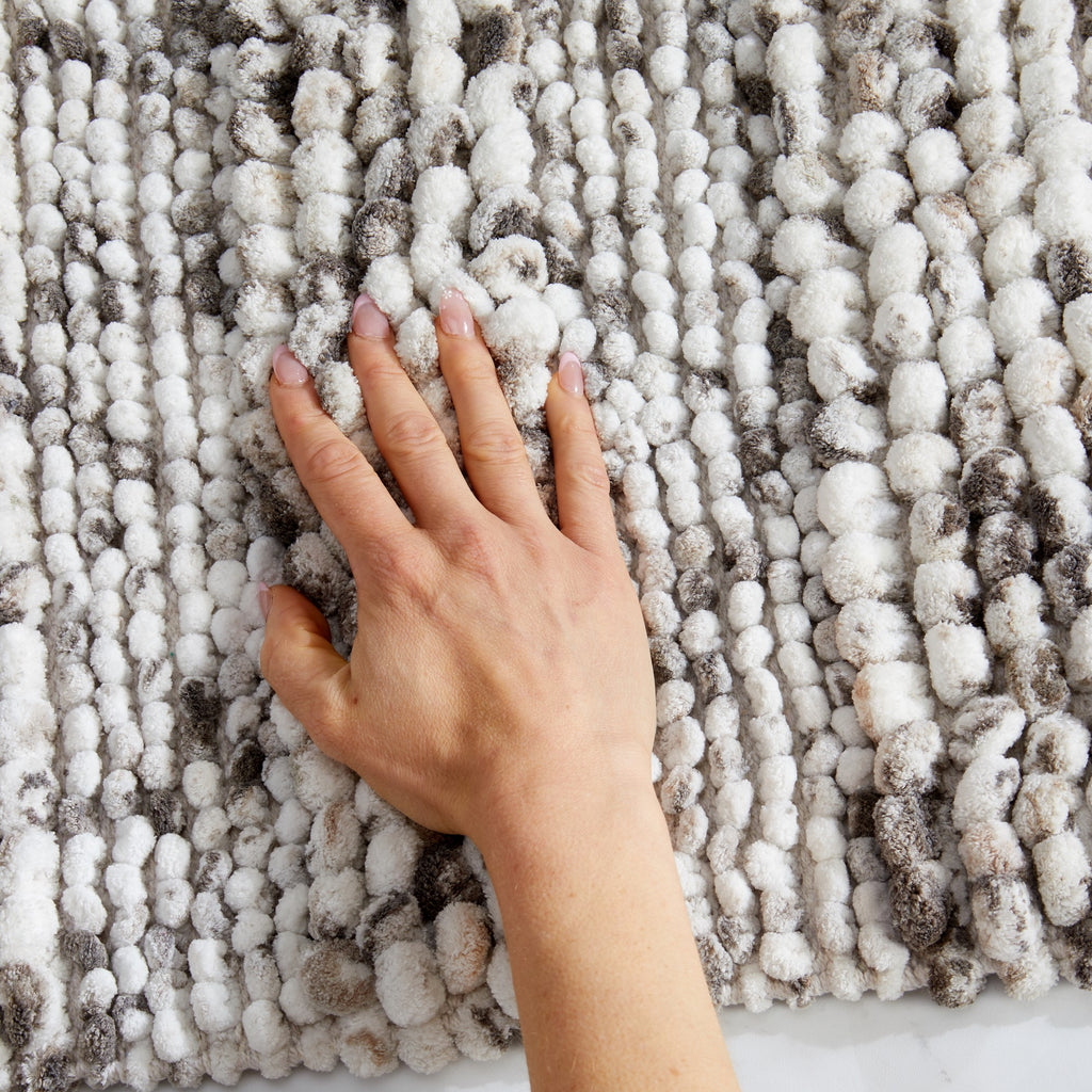 Close-up view of a space-dyed microfiber bath rug in ivory and gray tones with a hand resting on the surface to demonstrate its plush texture and softness. The thick, absorbent fibers provide cushioned comfort underfoot while adding visual texture and warmth to the bathroom.