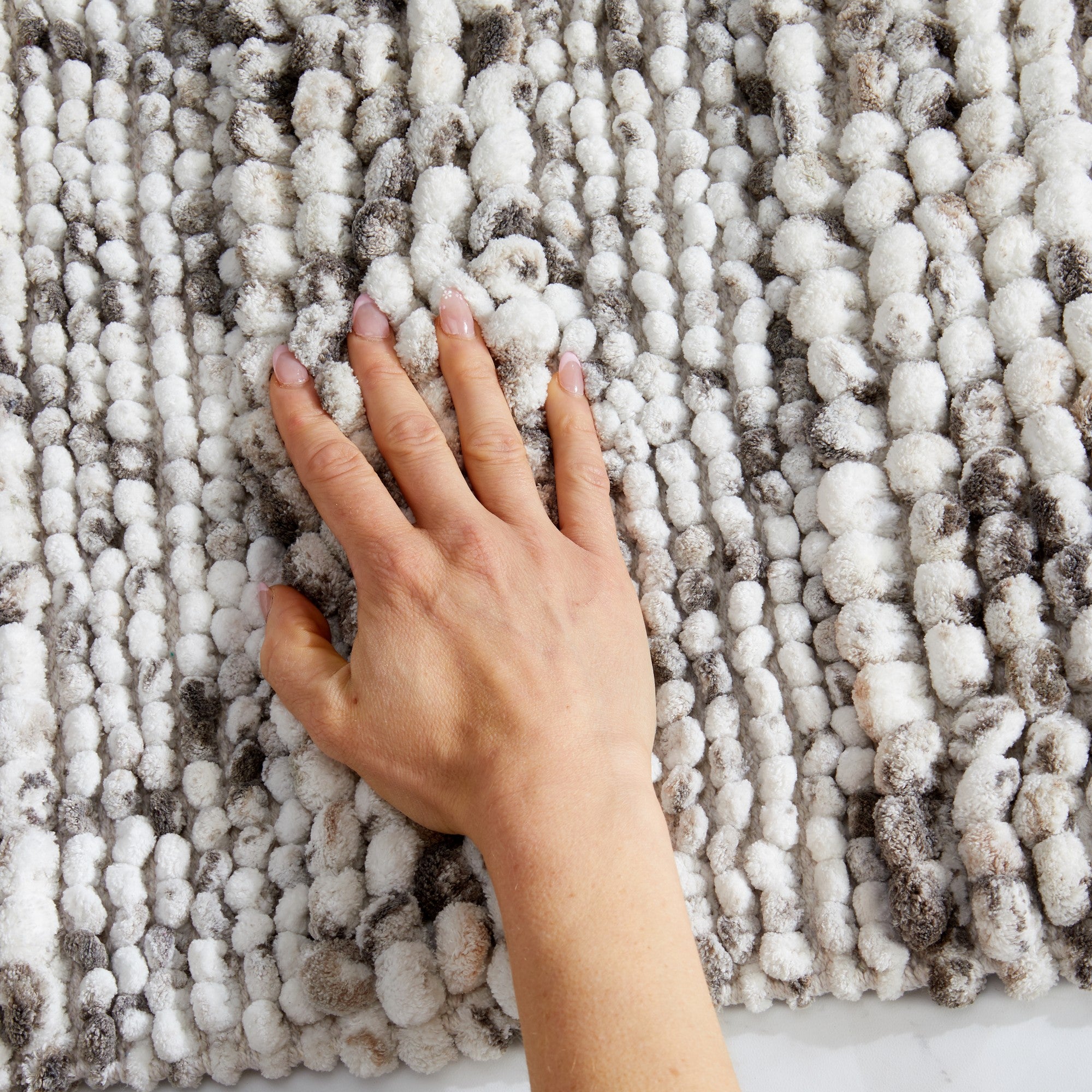 Close-up view of a space-dyed microfiber bath rug in ivory and gray tones with a hand resting on the surface to demonstrate its plush texture and softness. The thick, absorbent fibers provide cushioned comfort underfoot while adding visual texture and warmth to the bathroom.