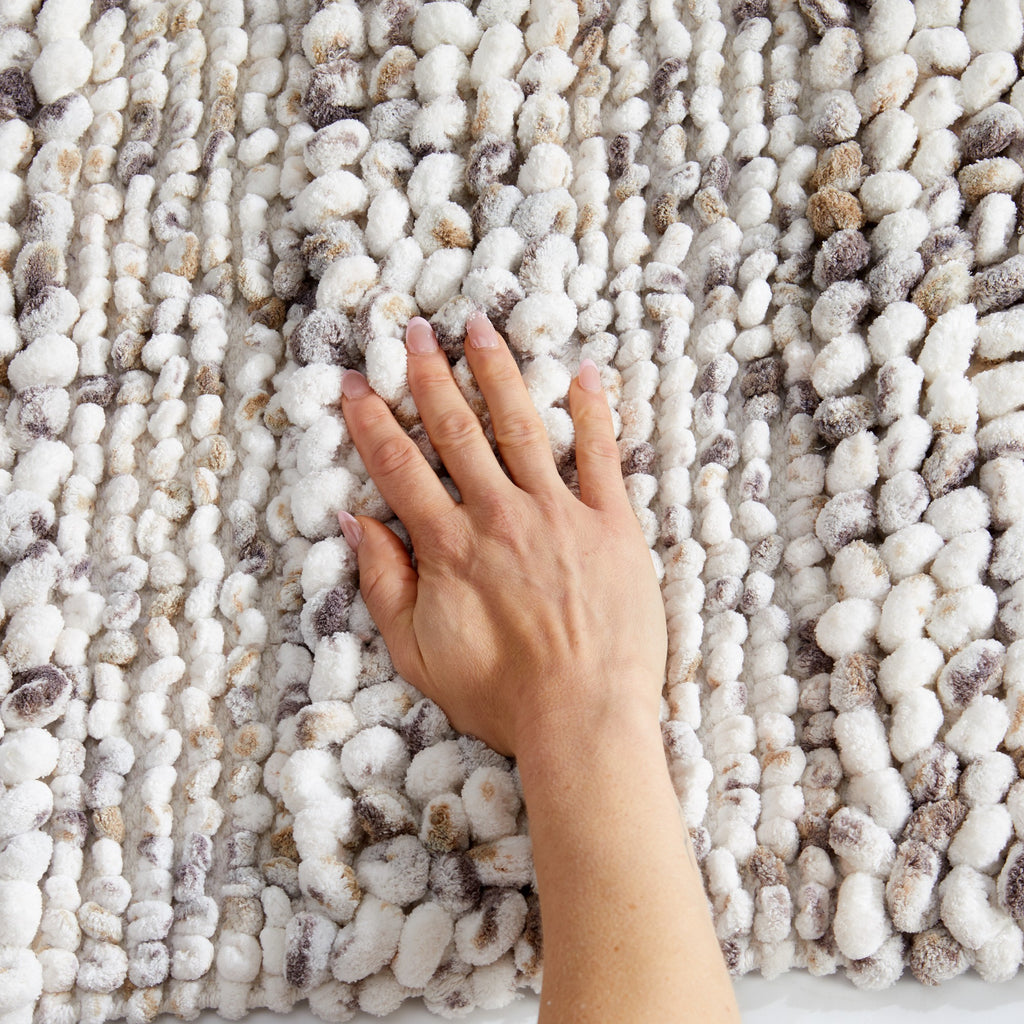 Close-up view of a plush space-dyed microfiber bath rug with a soft, textured pebble-like pile in ivory, gray, and beige tones. A hand presses into the surface to demonstrate the rug’s thick cushioning, softness, and comfort underfoot.