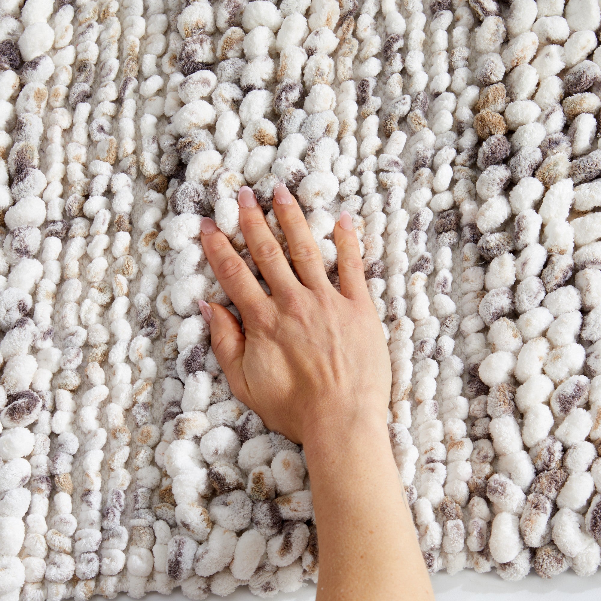 Close-up view of a plush space-dyed microfiber bath rug with a soft, textured pebble-like pile in ivory, gray, and beige tones. A hand presses into the surface to demonstrate the rug’s thick cushioning, softness, and comfort underfoot.