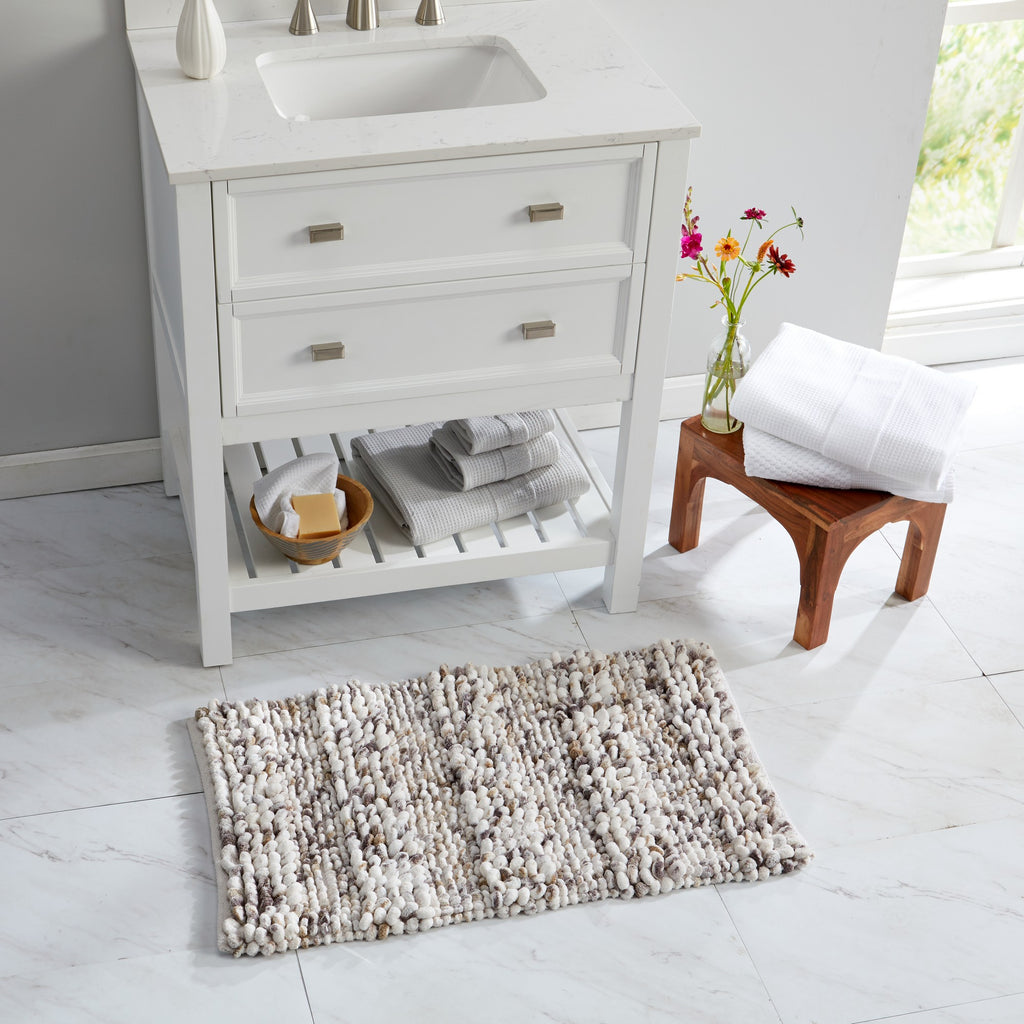 Space-dyed microfiber bath rug styled in a bright bathroom setting. The textured rug in soft ivory, gray, and beige tones is placed in front of a white vanity, showcasing its plush construction and versatile neutral design that complements modern bathroom décor.