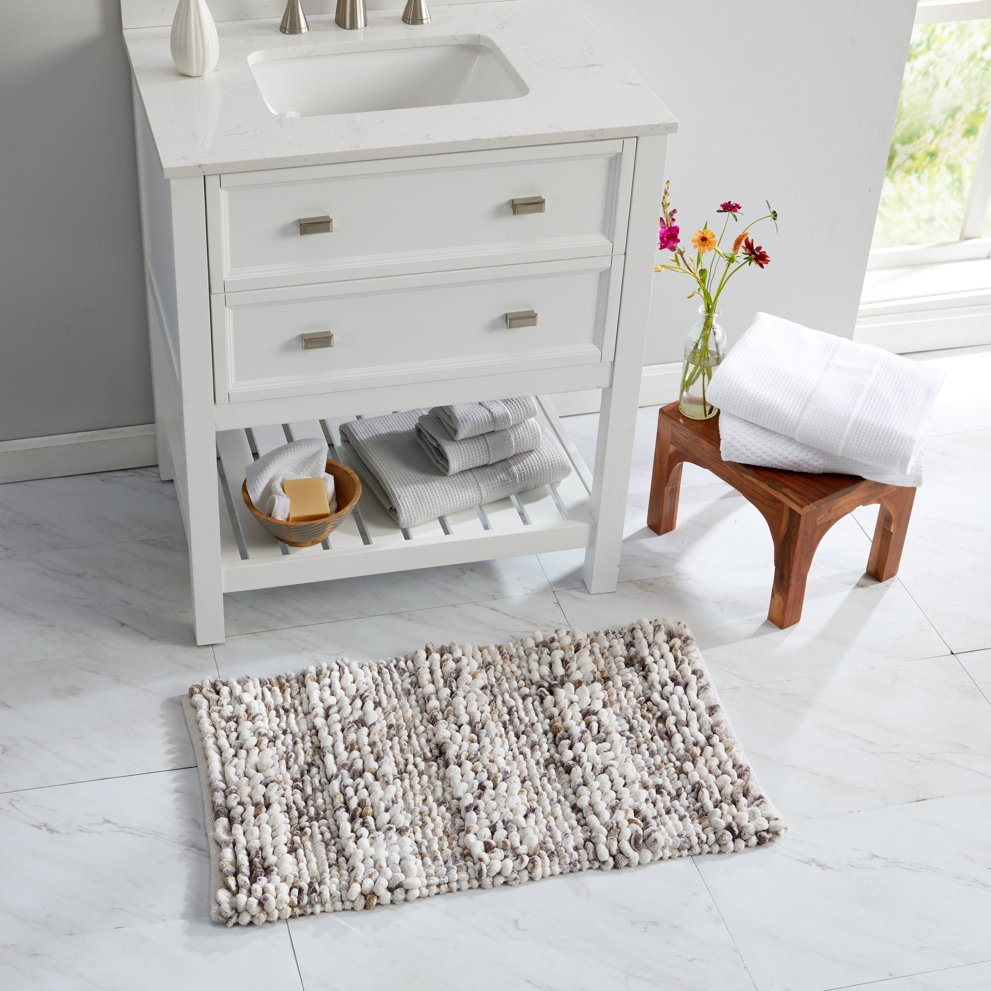 Space-dyed microfiber bath rug styled in a bright bathroom setting. The textured rug in soft ivory, gray, and beige tones is placed in front of a white vanity, showcasing its plush construction and versatile neutral design that complements modern bathroom décor.