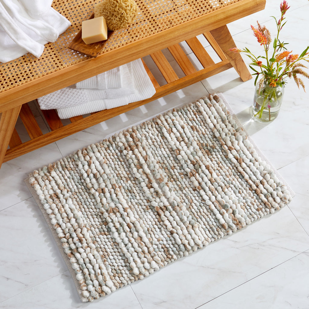 Space-dyed microfiber bath rug displayed in a bathroom setting beneath a wooden bench. The rug features a textured pebble-like design in soft ivory, beige, taupe, and light gray tones, offering plush comfort and a warm, natural look for any bathroom space.