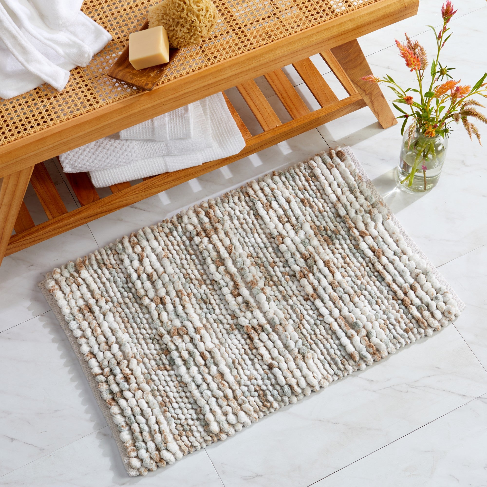 Space-dyed microfiber bath rug displayed in a bathroom setting beneath a wooden bench. The rug features a textured pebble-like design in soft ivory, beige, taupe, and light gray tones, offering plush comfort and a warm, natural look for any bathroom space.