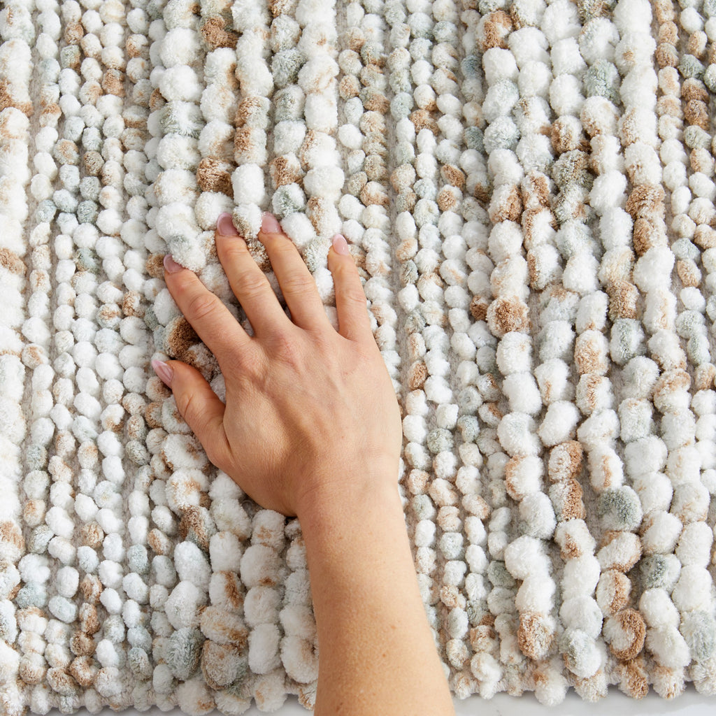 Close-up view of a space-dyed microfiber bath rug featuring a plush pebble-textured surface in ivory, beige, taupe, and soft gray tones. A hand presses into the thick pile to demonstrate the rug’s softness, cushioning, and comfortable feel underfoot.