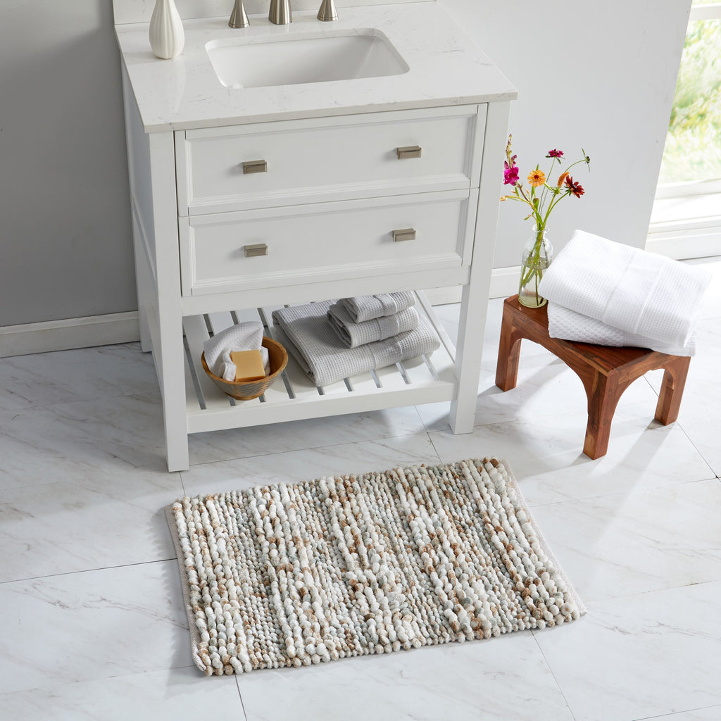 Space-dyed microfiber bath rug displayed in a bright bathroom setting in front of a white vanity. The textured pebble-like surface features a blend of ivory, beige, taupe, and soft gray tones, offering plush comfort and a versatile neutral design that complements a variety of bathroom décor styles.