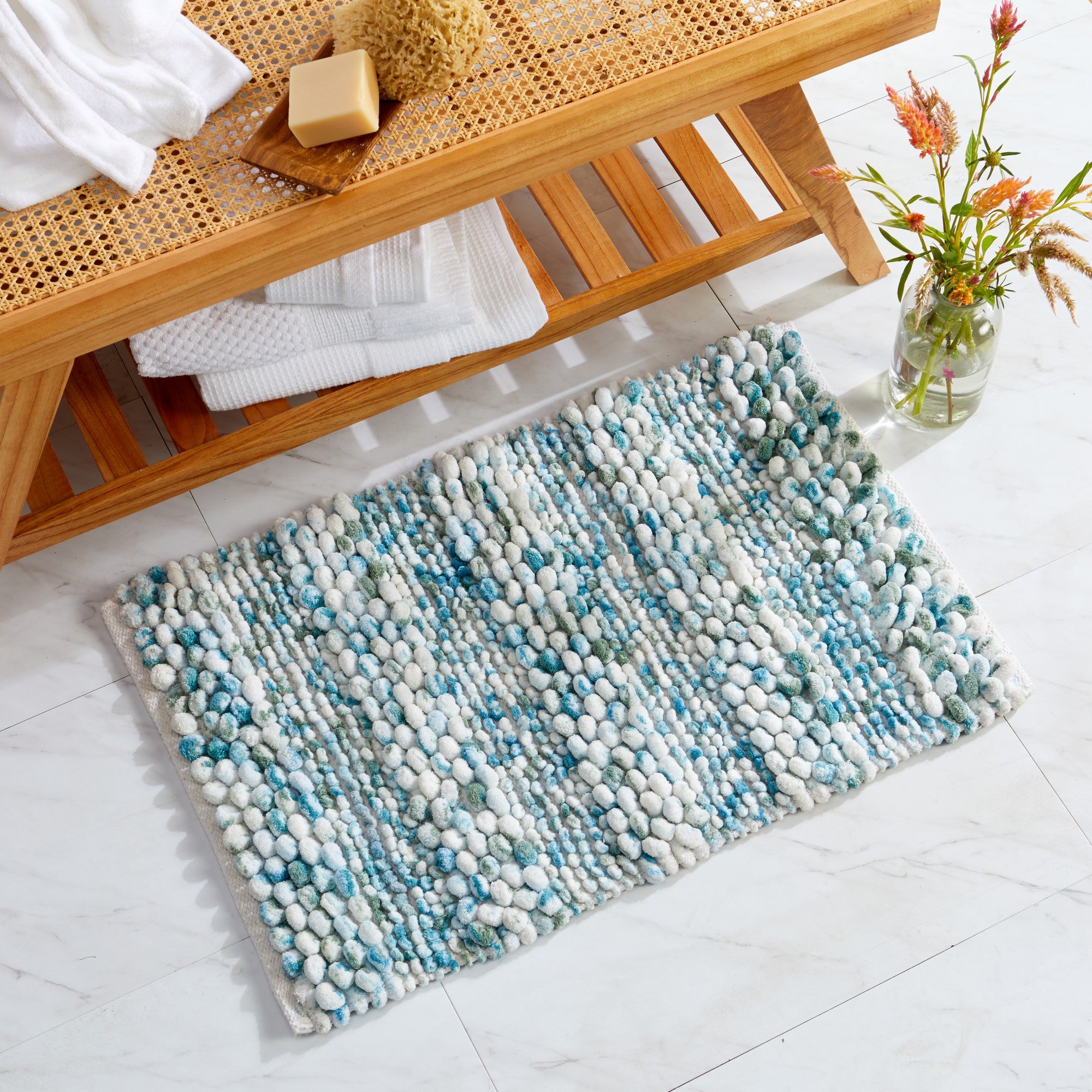 Space-dyed microfiber bath rug displayed in a bathroom setting beneath a wooden bench. The rug features a plush pebble-textured design in white, aqua blue, turquoise, and soft gray tones, bringing a fresh coastal-inspired look while providing soft comfort underfoot.