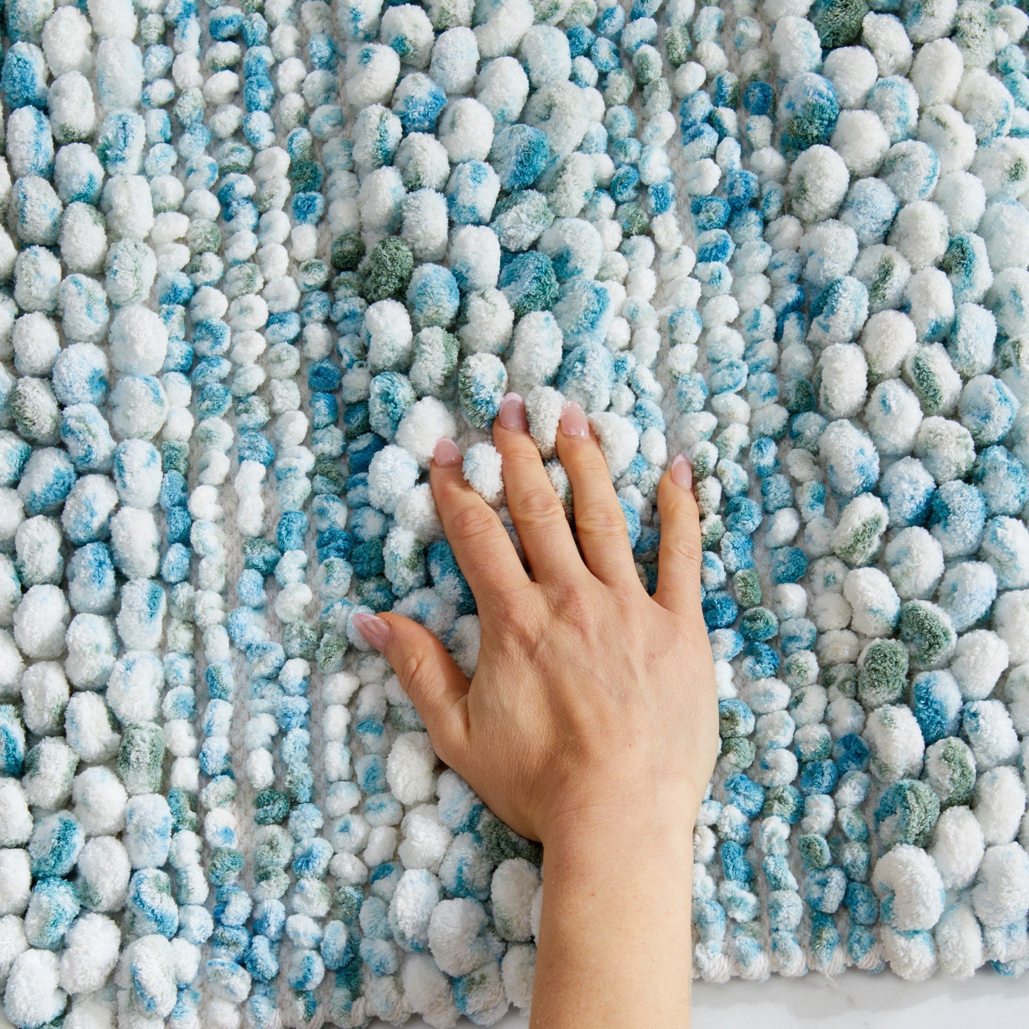 Close-up view of a plush space-dyed microfiber bath rug featuring a pebble-textured surface in white, aqua blue, turquoise, and soft gray tones. A hand presses into the thick pile to demonstrate the rug’s softness, cushioning, and luxurious comfort underfoot.