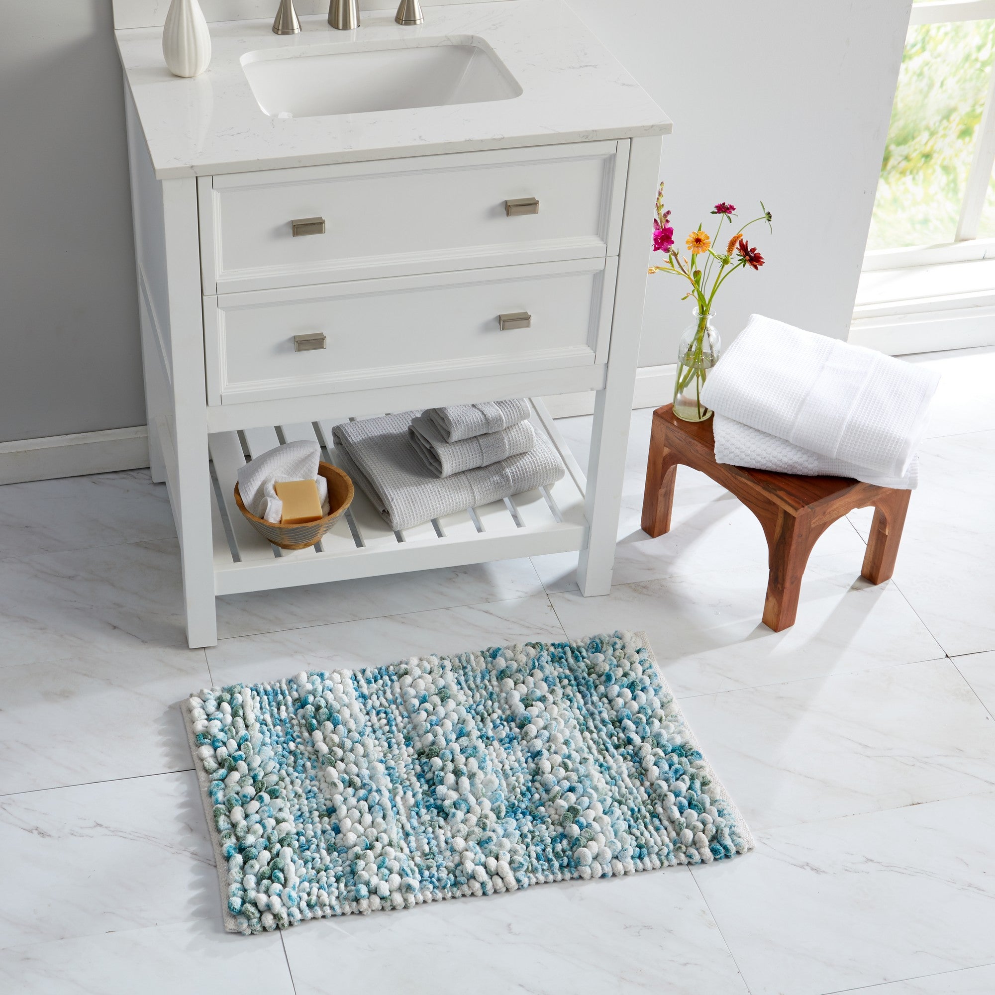 Space-dyed microfiber bath rug displayed in a bright bathroom setting in front of a white vanity. The plush pebble-textured surface features a blend of white, aqua blue, turquoise, and soft gray tones, adding a fresh coastal-inspired look while providing soft comfort and cushioning underfoot.