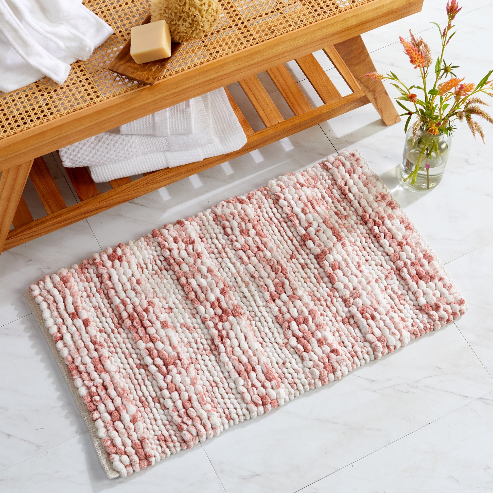 Space-dyed microfiber bath rug featuring a plush pebble-textured design in soft blush pink and ivory tones. Displayed in a bright bathroom setting beside a wooden bench, the rug provides ultra-soft comfort, absorbency, and a warm, inviting accent for any bathroom décor.