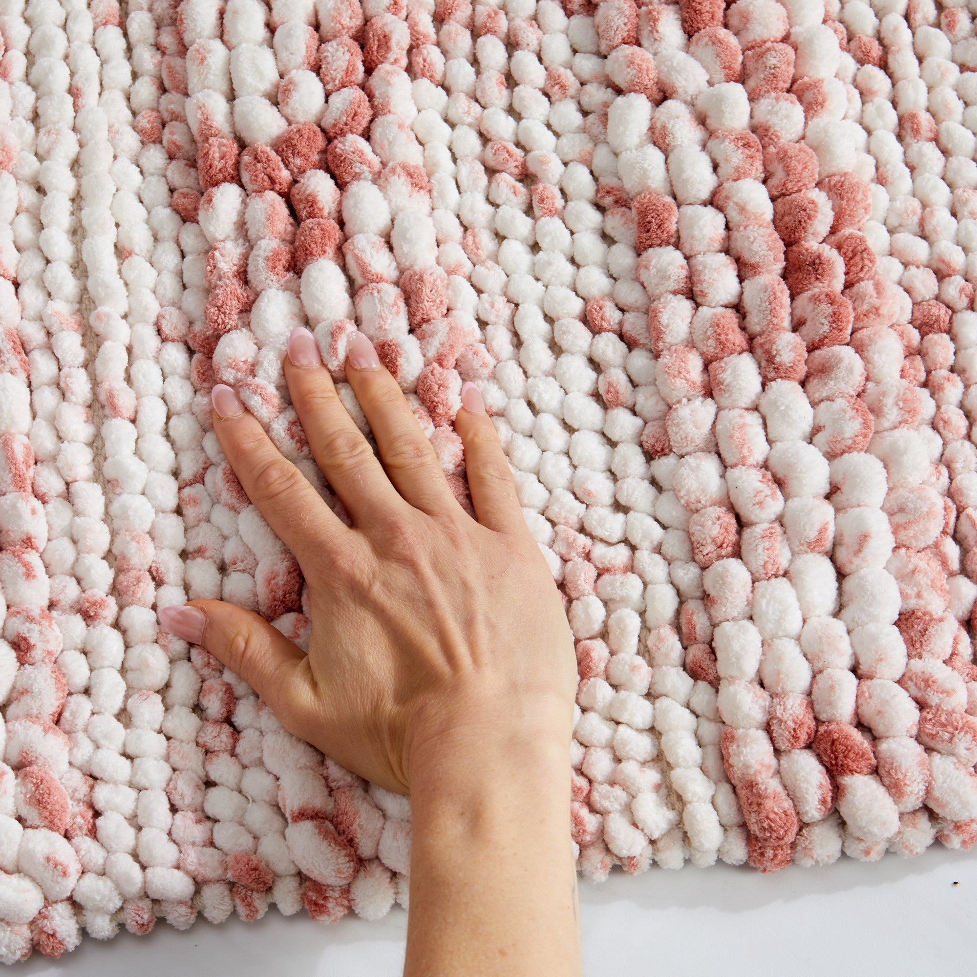 Close-up view of a plush space-dyed microfiber bath rug in soft blush pink and ivory tones. A hand presses into the thick pebble-textured surface, highlighting the rug's ultra-soft feel, dense pile, and cushioned comfort underfoot.