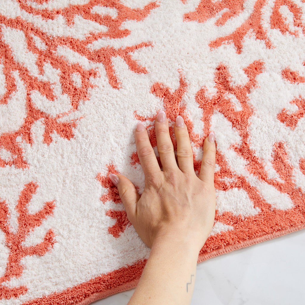 Close-up view of a coastal-inspired bath rug featuring a coral branch pattern in soft coral pink on an ivory background. A hand rests on the plush surface, highlighting the rug's soft texture, cushioned feel, and comfortable underfoot support.