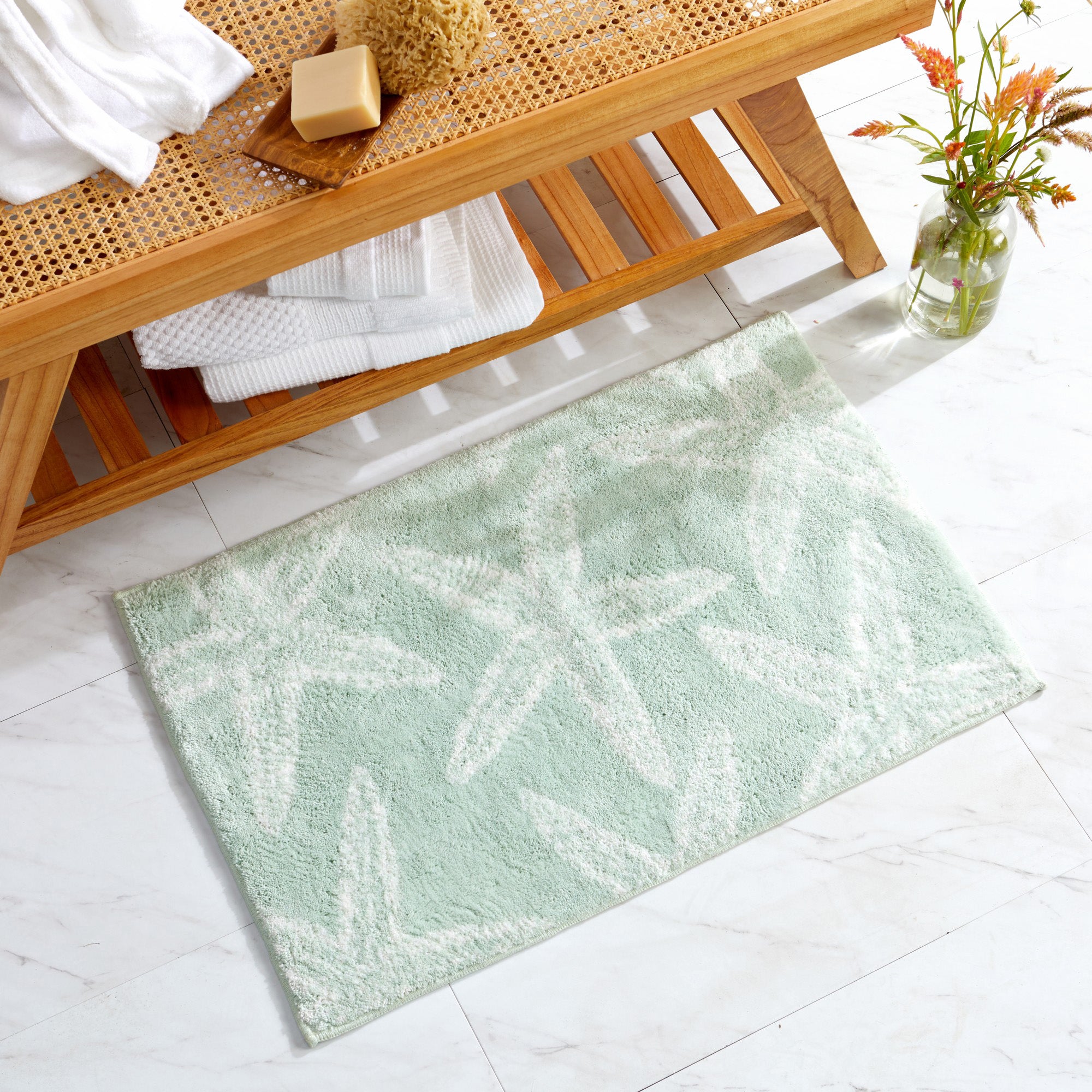 Coastal-themed bath rug featuring a soft seafoam green background with white starfish motifs. Displayed in a bright bathroom setting, the plush and absorbent rug adds a refreshing beach-inspired accent while providing comfort and softness underfoot.