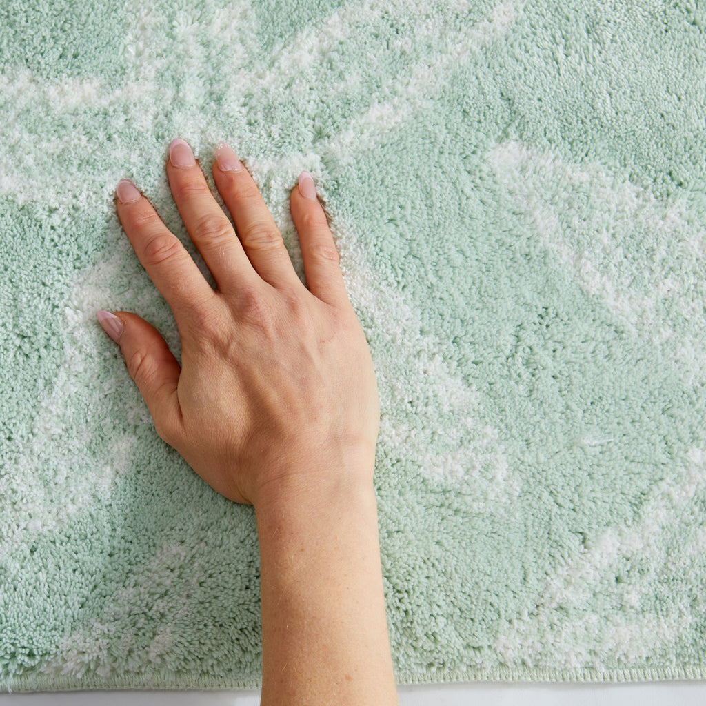Close-up view of a coastal-themed bath rug in soft seafoam green with white starfish motifs. A hand rests on the plush surface, highlighting the rug's soft texture, cushioned feel, and comfortable underfoot support. Ideal for adding a fresh, beach-inspired touch to bathroom décor.