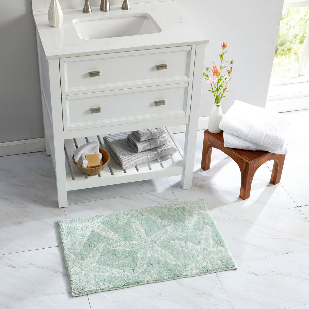 Coastal-themed bath rug featuring white starfish motifs on a soft seafoam green background, styled in a bright bathroom setting. The plush, absorbent rug adds a fresh beach-inspired accent while providing comfort, softness, and everyday functionality.