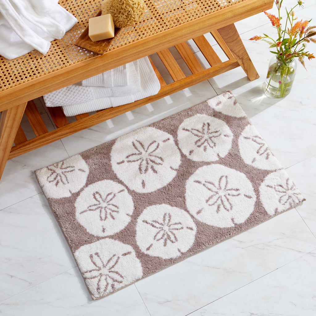 Coastal-inspired bath rug featuring oversized sand dollar motifs in soft ivory on a taupe background. The plush, absorbent design adds beach-inspired charm and comfort to bathrooms while providing a soft, cozy surface underfoot.