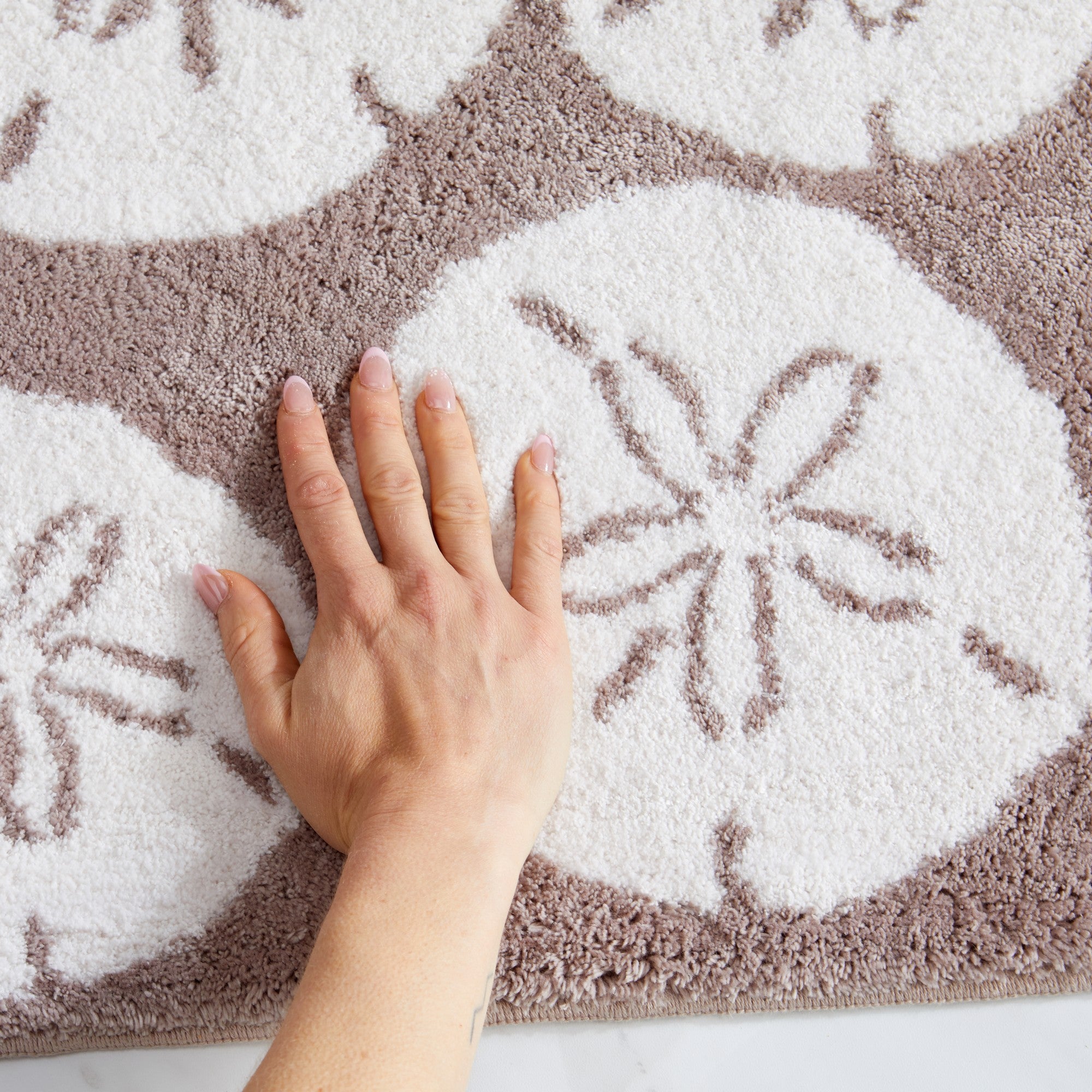 Close-up view of a plush coastal bath rug featuring oversized ivory sand dollar motifs on a soft taupe background. The image highlights the rug’s thick, cushioned texture and soft feel, demonstrating its comfort and absorbent construction.