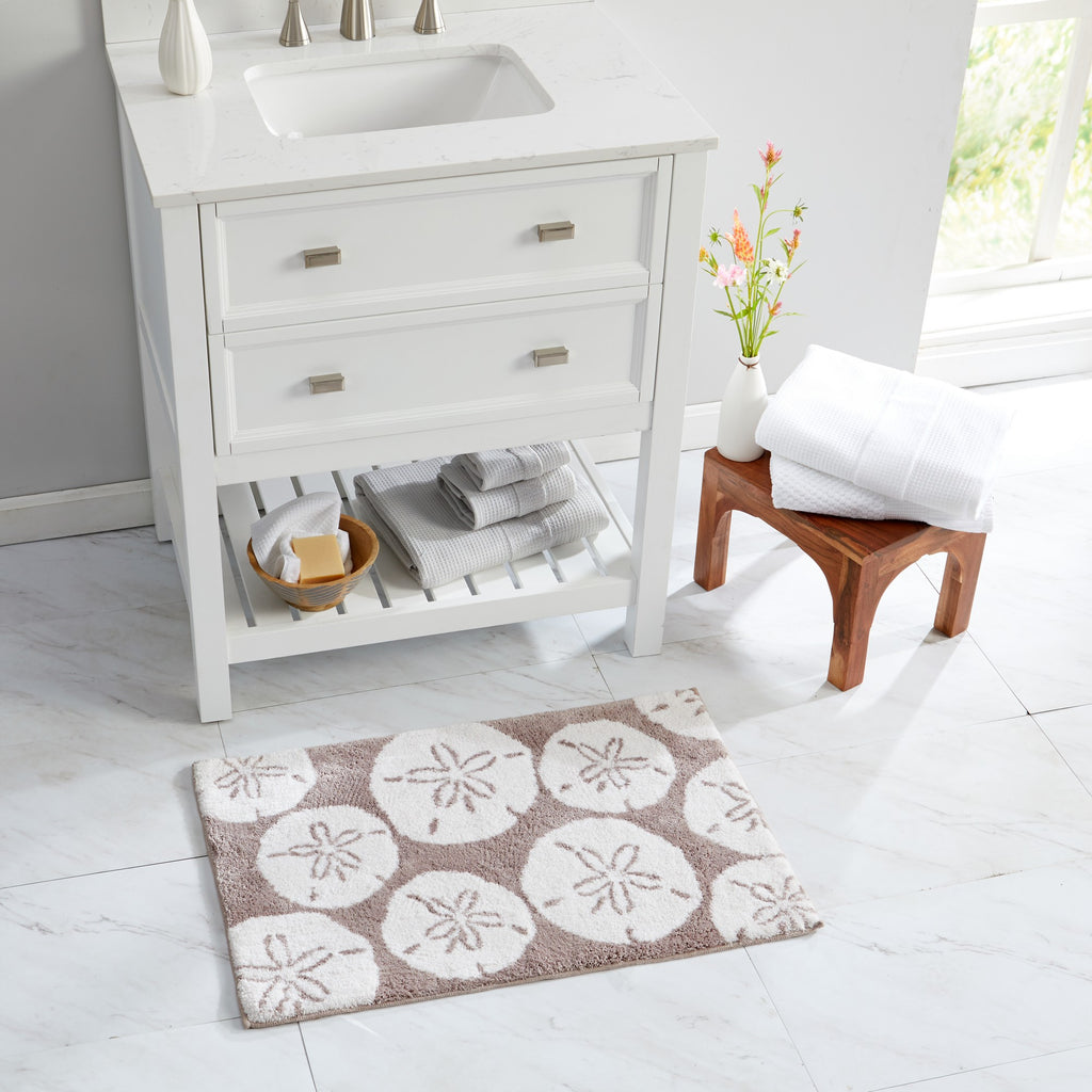 Coastal-inspired bath rug featuring oversized ivory sand dollar motifs on a soft taupe background, styled in a bright bathroom setting. The plush, absorbent rug adds beach-inspired charm, comfort, and texture to bathroom décor.