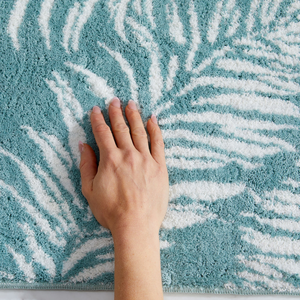 Close-up of a plush tropical bath rug featuring white palm leaf silhouettes on a soft aqua blue background. The image highlights the rug’s thick, cushioned texture and absorbent pile, providing comfort and a fresh coastal-inspired look.