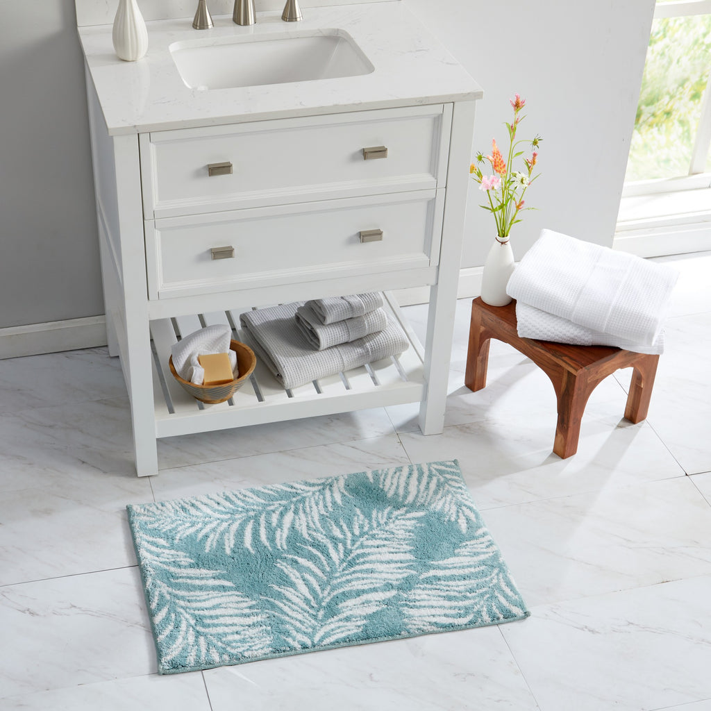 Bathroom scene featuring a plush tropical bath rug with white palm leaf silhouettes on an aqua blue background. The rug is displayed in front of a white vanity with folded towels and natural wood accents, showcasing its coastal-inspired style and soft, comfortable texture.