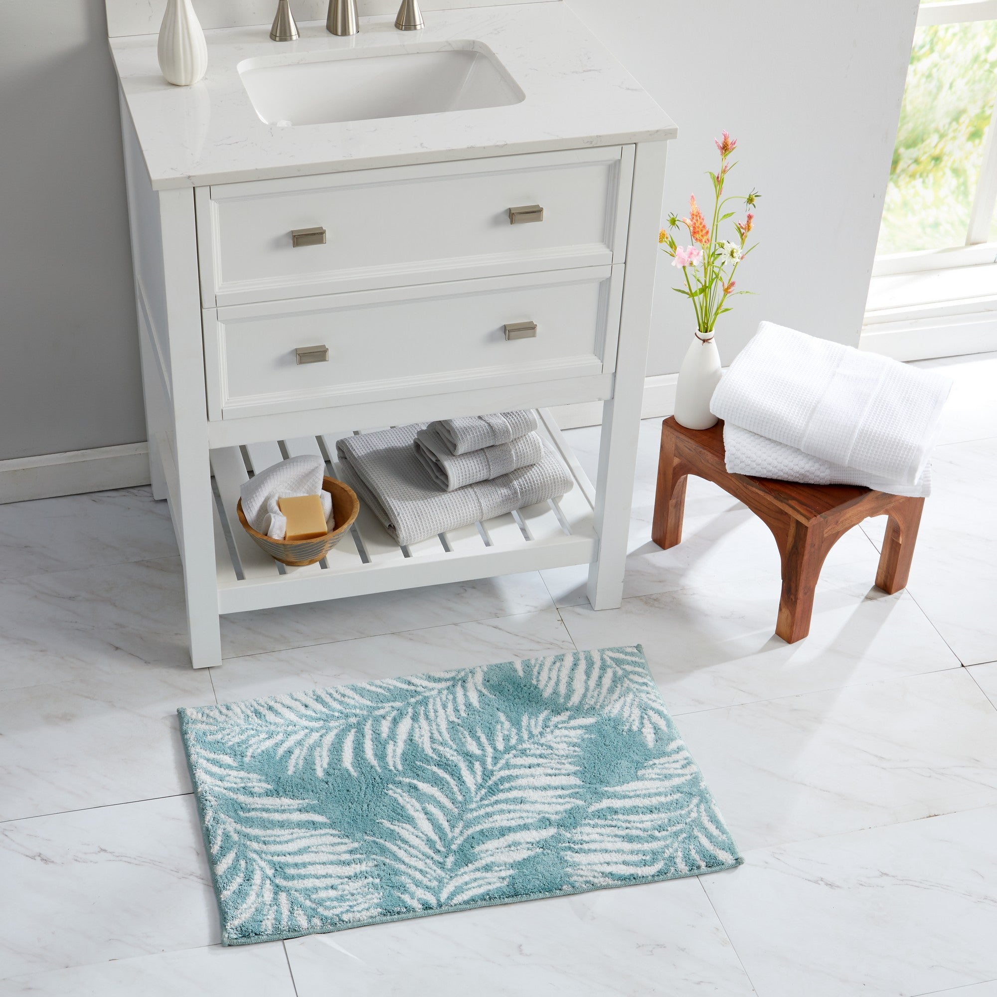 Bathroom scene featuring a plush tropical bath rug with white palm leaf silhouettes on an aqua blue background. The rug is displayed in front of a white vanity with folded towels and natural wood accents, showcasing its coastal-inspired style and soft, comfortable texture.