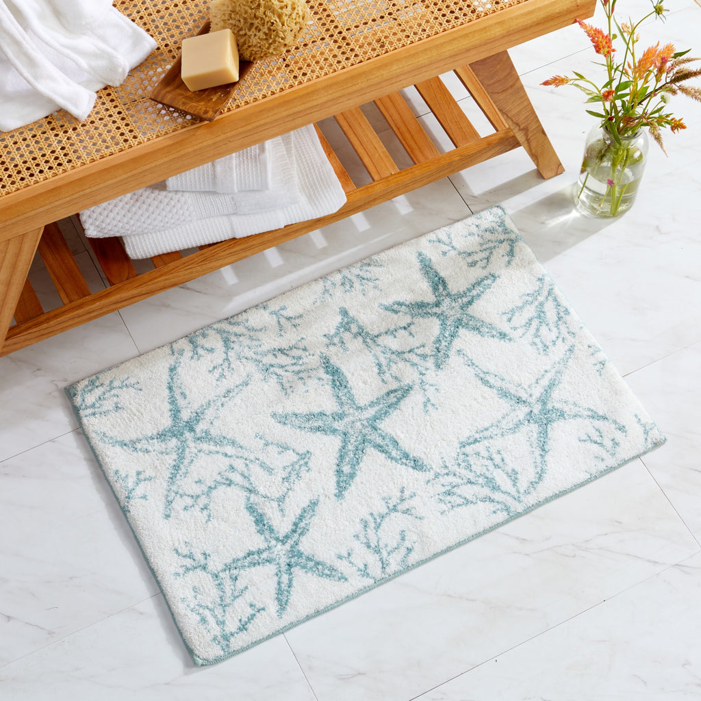 Coastal-themed bath rug featuring aqua blue starfish and coral motifs on a soft white background. Displayed in a bright bathroom setting with natural wood accents and folded towels, the plush rug adds a fresh seaside touch while providing comfort underfoot.