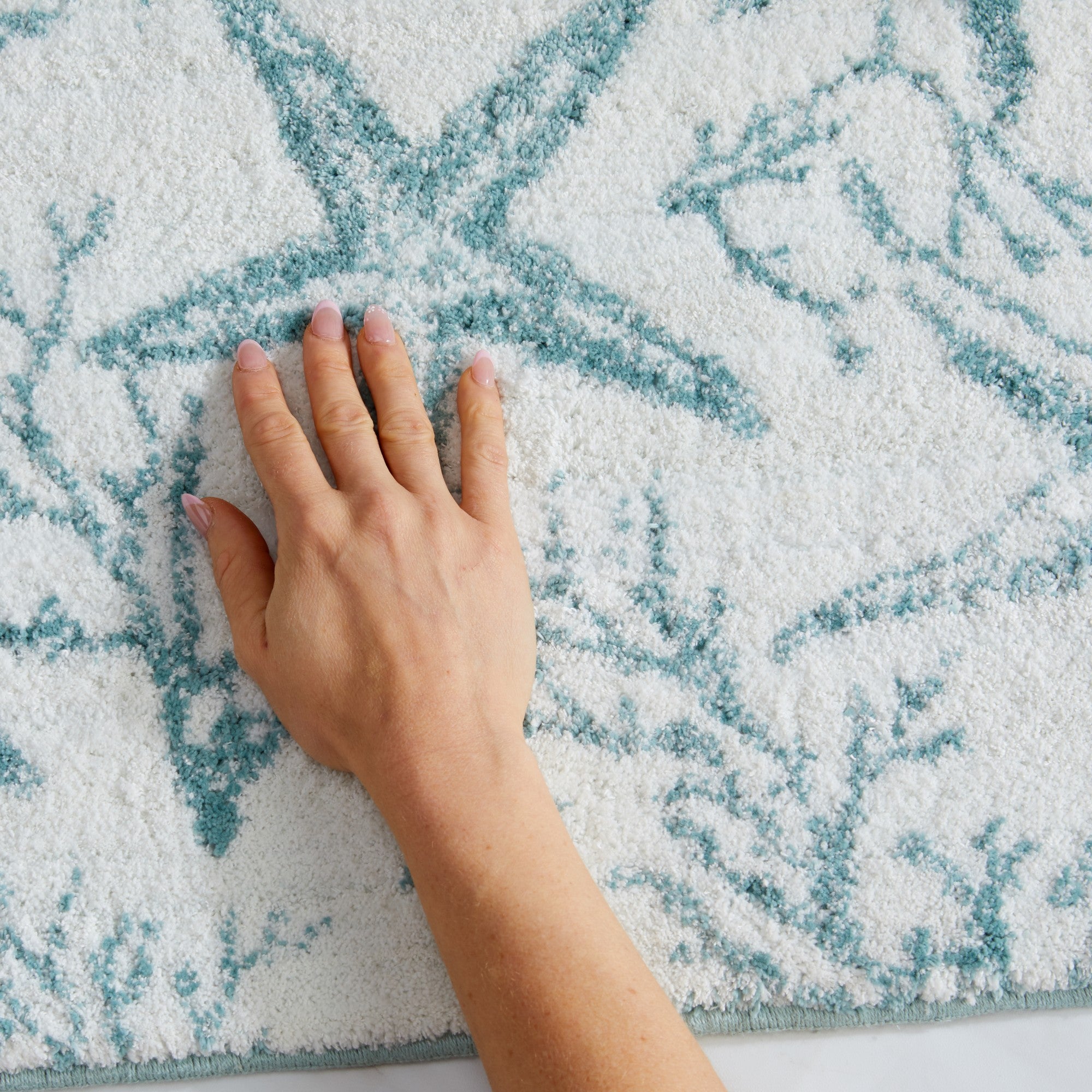 Close-up of a plush coastal bath rug featuring aqua blue starfish and coral branch patterns on a soft white background. A hand presses into the textured surface to showcase the rug’s thick pile, softness, and cushioned feel underfoot.