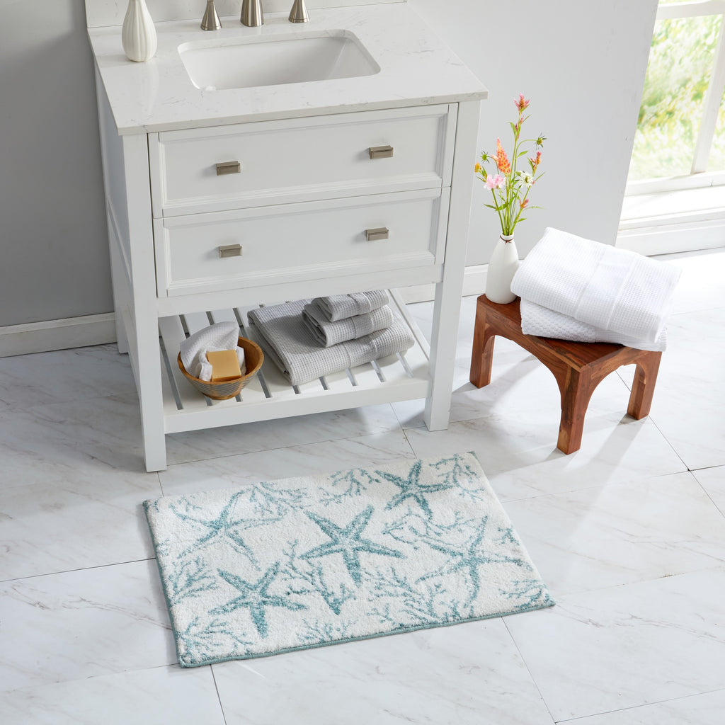 Coastal-inspired bath rug featuring aqua blue starfish and coral patterns on a soft white background. Styled in a bright bathroom beside a white vanity and natural wood stool, the plush rug adds seaside charm while providing comfort and absorbency for everyday use.