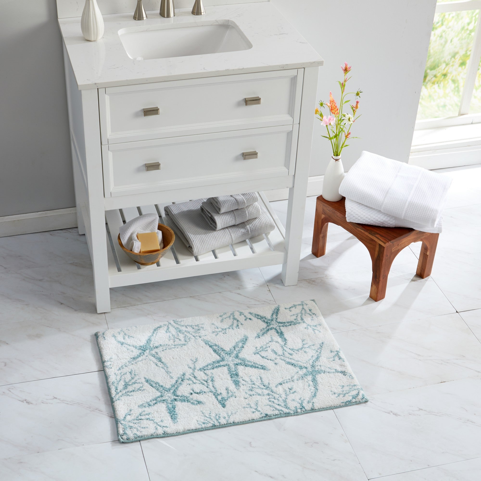 Coastal-inspired bath rug featuring aqua blue starfish and coral patterns on a soft white background. Styled in a bright bathroom beside a white vanity and natural wood stool, the plush rug adds seaside charm while providing comfort and absorbency for everyday use.