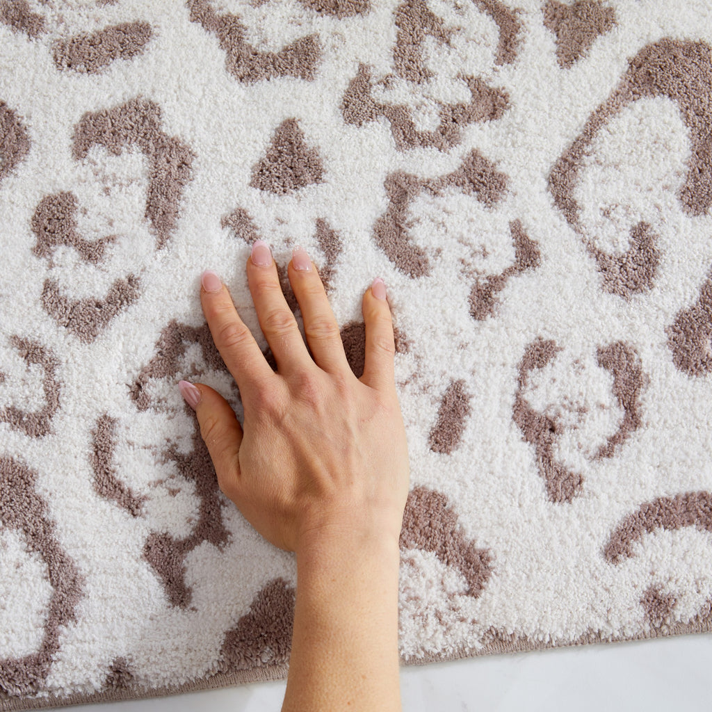 Close-up of a plush bath rug featuring a blush pink leopard print pattern on a soft ivory background. A hand presses into the thick, textured pile to showcase the rug’s softness, cushioning, and comfortable feel underfoot.