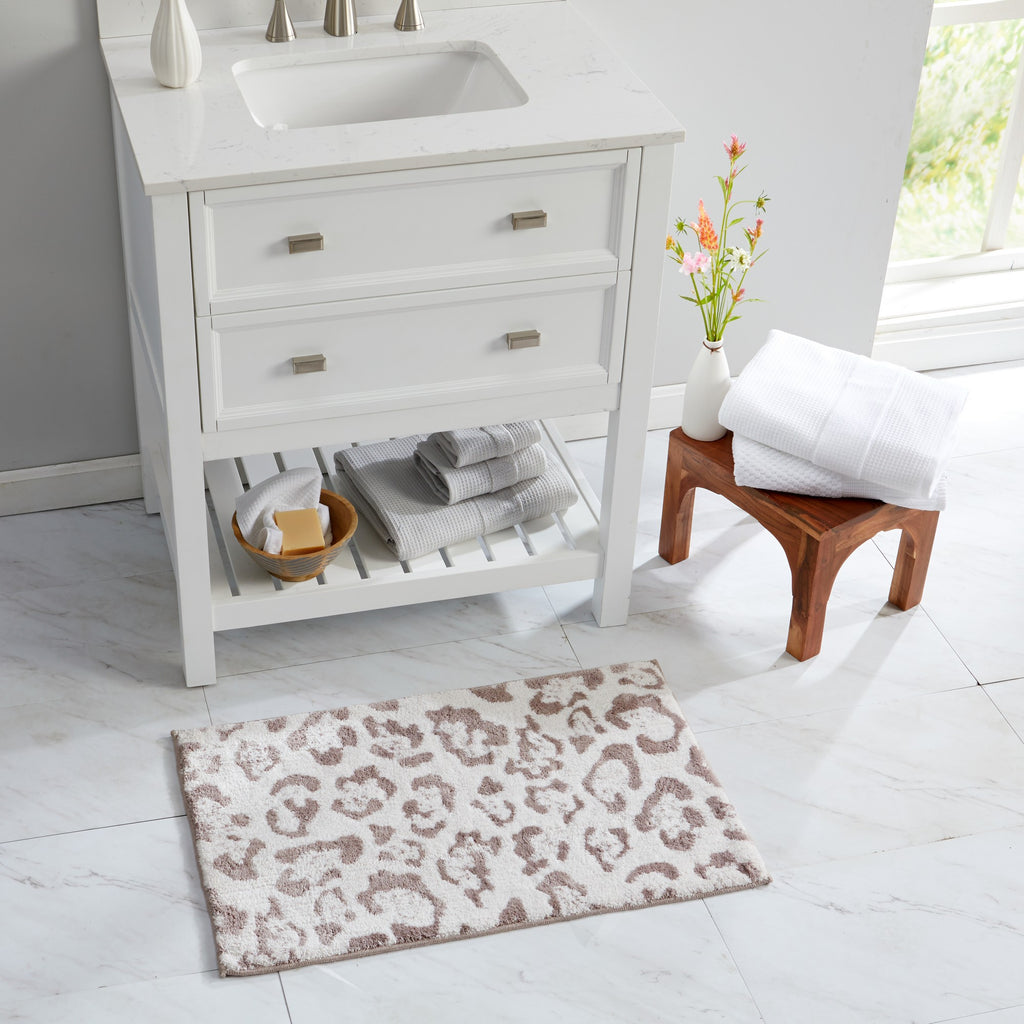 Close-up of a plush bath rug featuring a blush pink leopard print pattern on a soft ivory background. A hand presses into the thick, textured pile to showcase the rug’s softness, cushioning, and comfortable feel underfoot.