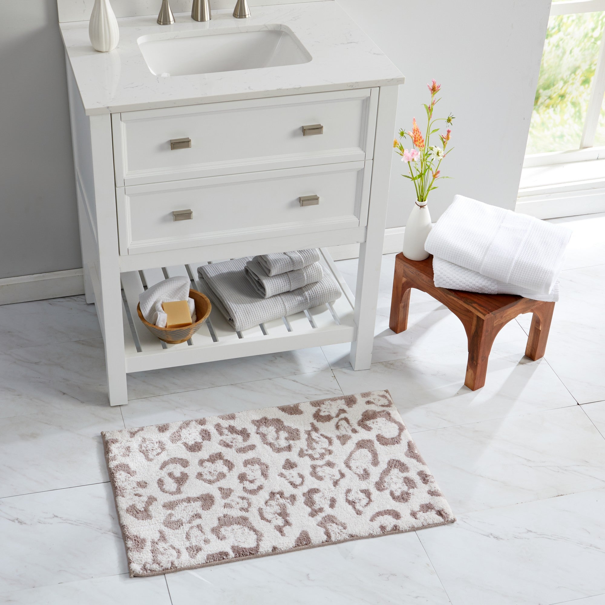 Close-up of a plush bath rug featuring a blush pink leopard print pattern on a soft ivory background. A hand presses into the thick, textured pile to showcase the rug’s softness, cushioning, and comfortable feel underfoot.