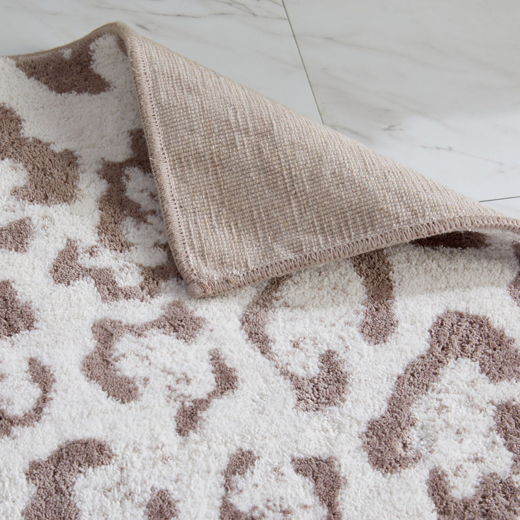 Close-up of a plush leopard-print bath rug with a blush pink pattern on an ivory background. One corner is folded back to reveal the durable woven backing, highlighting the rug’s construction, texture, and quality craftsmanship.