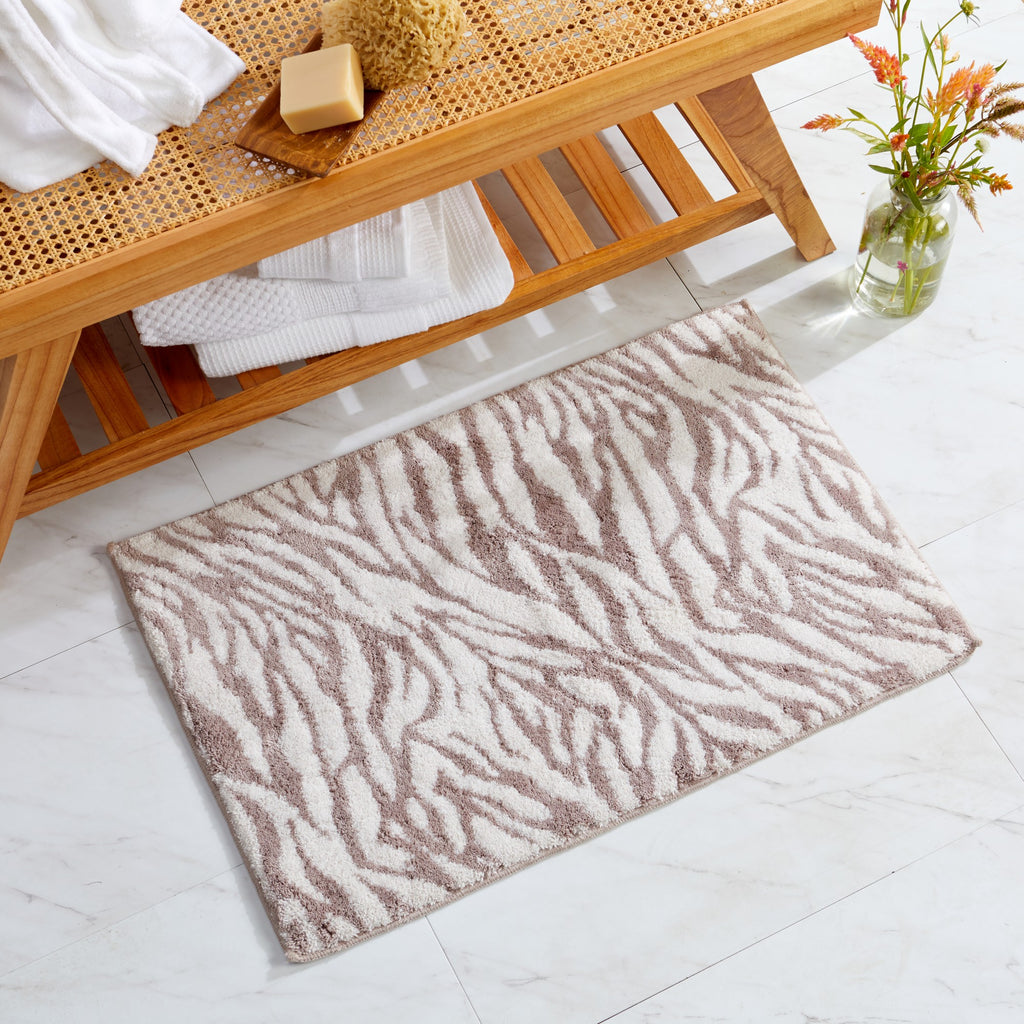 Animal-inspired bath rug featuring a taupe zebra stripe pattern on a soft ivory background. Styled in a bright bathroom setting with natural wood accents, the plush rug adds a modern decorative touch while providing comfort, absorbency, and softness underfoot.