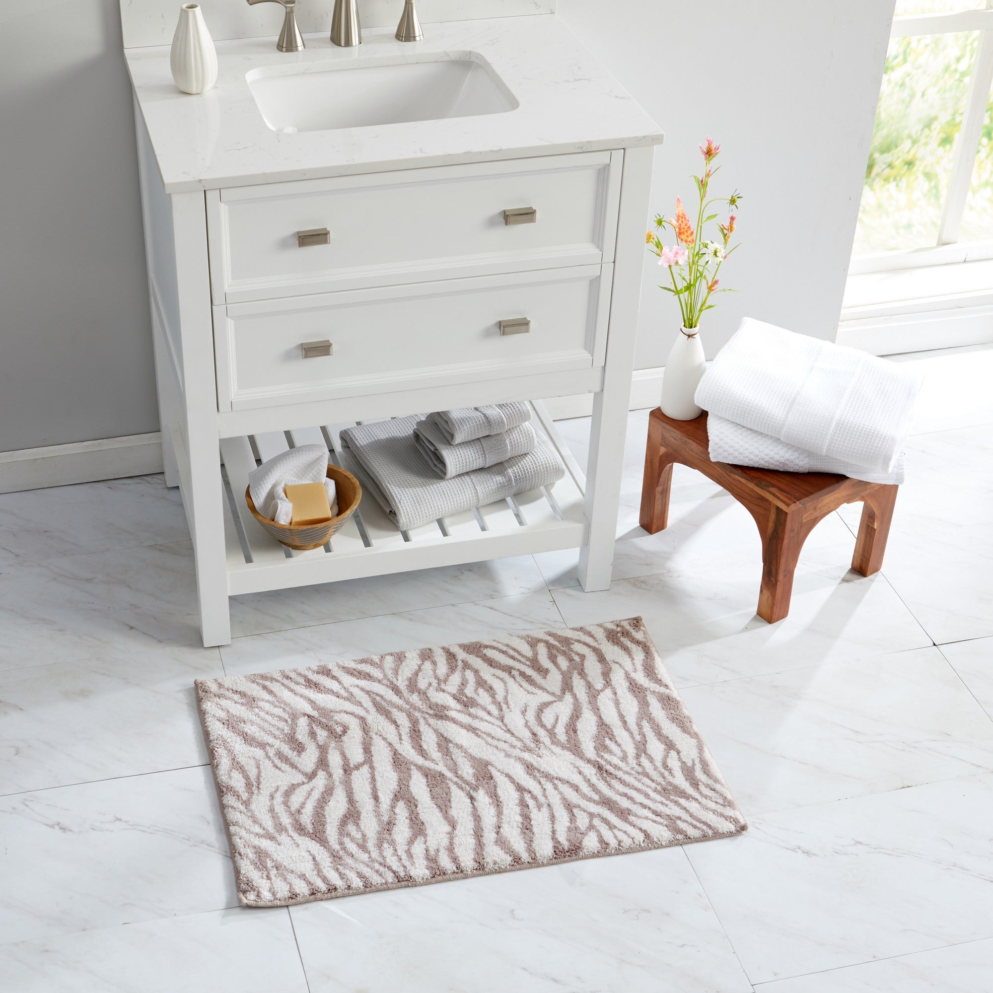 Taupe and ivory zebra-print bath rug styled in a bright bathroom setting beside a white vanity. The plush rug adds a modern animal-inspired accent while providing soft comfort underfoot, with neatly folded towels and natural wood décor completing the spa-like look.