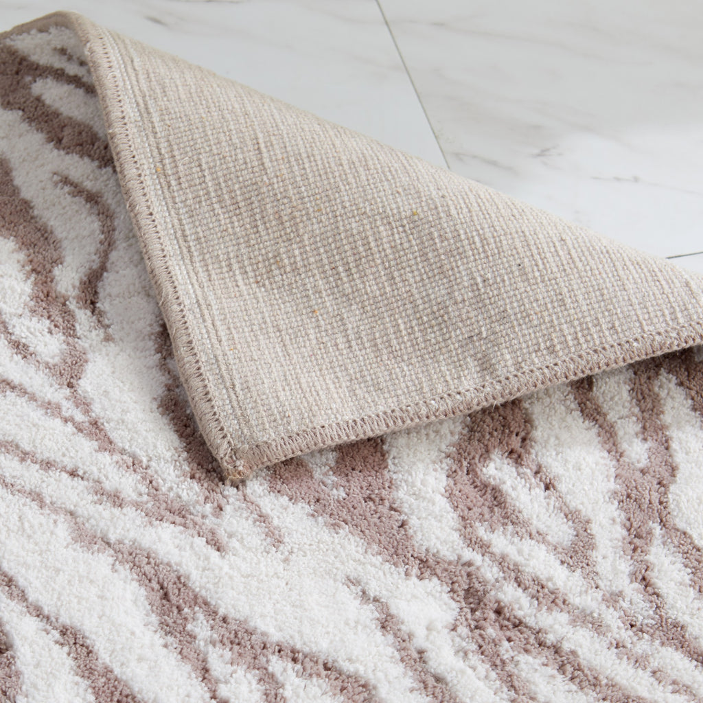 Close-up view of a taupe and ivory zebra-print bath rug with a lifted corner showcasing the durable woven backing. The plush, textured surface highlights the rug’s soft feel, quality construction, and stylish animal-inspired pattern.