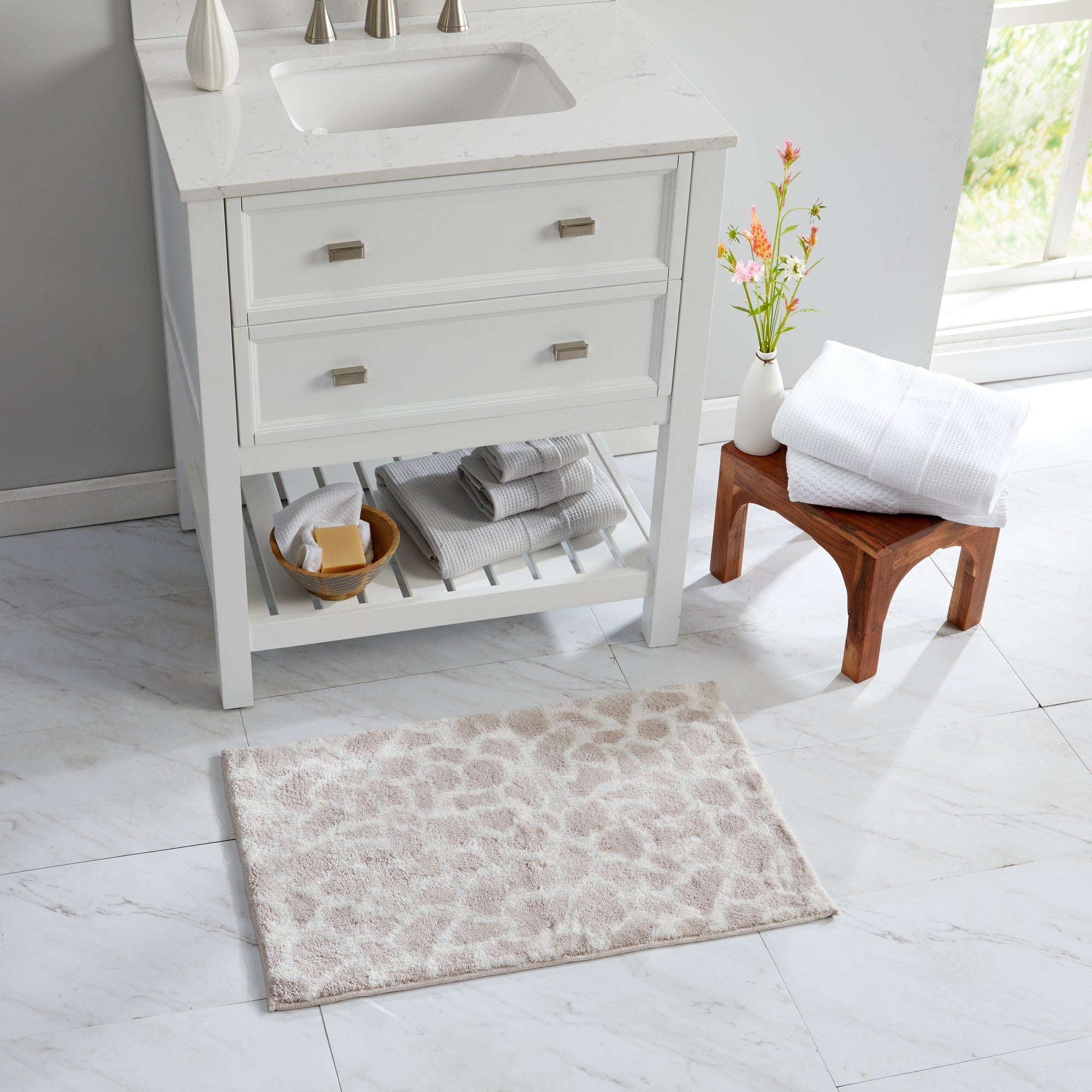 Giraffe-print bath rug styled in a bright bathroom beside a white vanity and neatly folded towels. The soft ivory and taupe pattern adds a subtle safari-inspired accent while providing plush comfort and absorbent performance for everyday use.