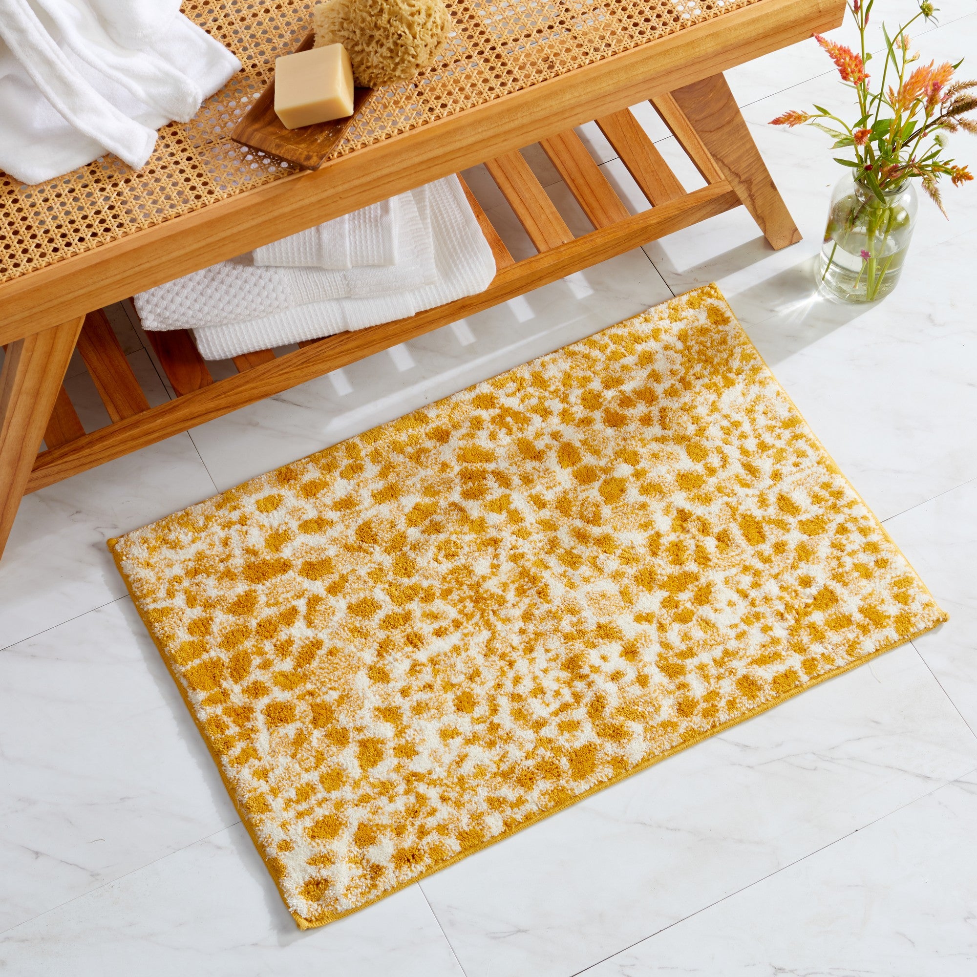 Mustard yellow and ivory cheetah-print bath rug displayed in a spa-inspired bathroom setting beside a natural wood bench and decorative vase. The plush, textured rug adds a bold animal-print accent while providing soft, absorbent comfort for everyday use.