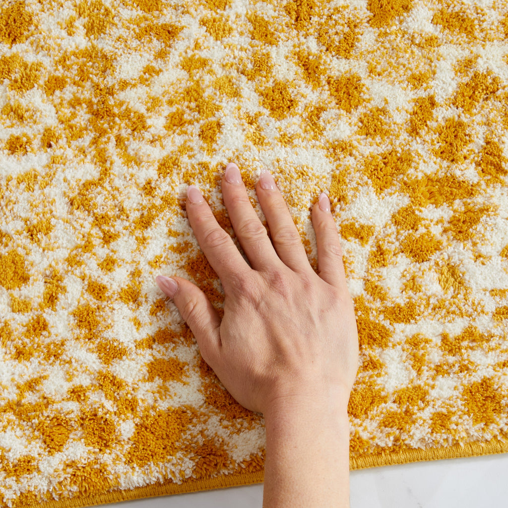 Close-up of a mustard yellow and ivory cheetah-print bath rug with a hand resting on the surface to demonstrate its plush texture and softness. The thick pile highlights the rug’s cushioned feel, absorbency, and comfortable underfoot support for everyday bathroom use.