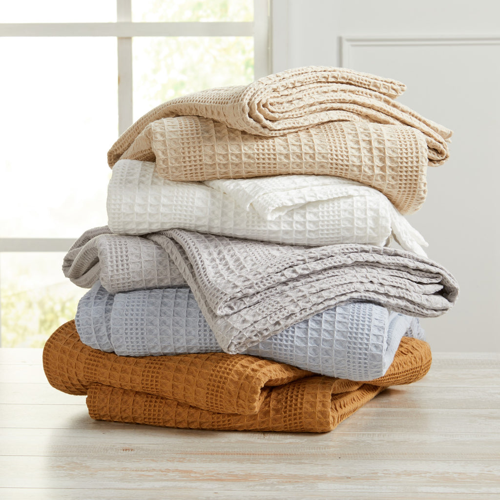 A stack of various sized cotton waffle weave blankets in beige, white, gray, and orange, neatly folded and placed on a wooden surface with a window in the background.