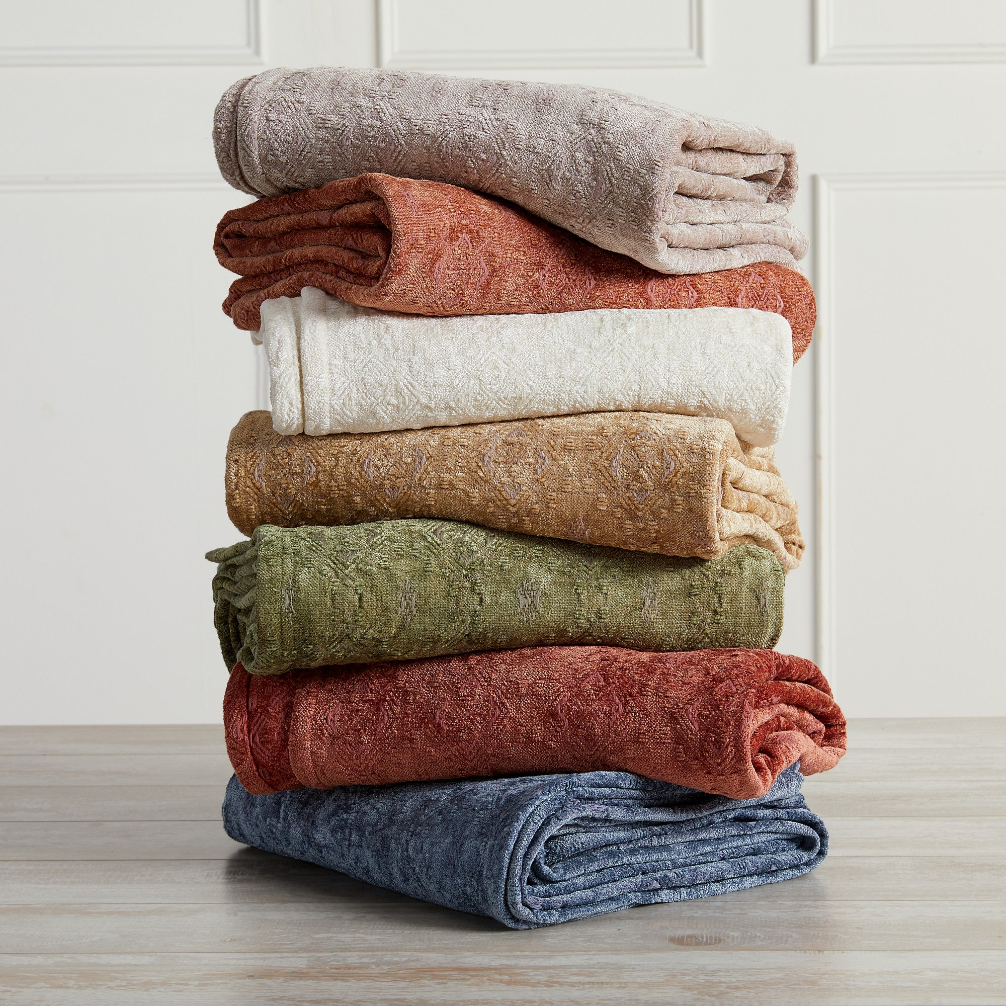 A stack of folded chenille jacquard throw blankets in various solid colors including blue, gray, rose, sage, clay, beige, and white, placed on a wooden surface.
