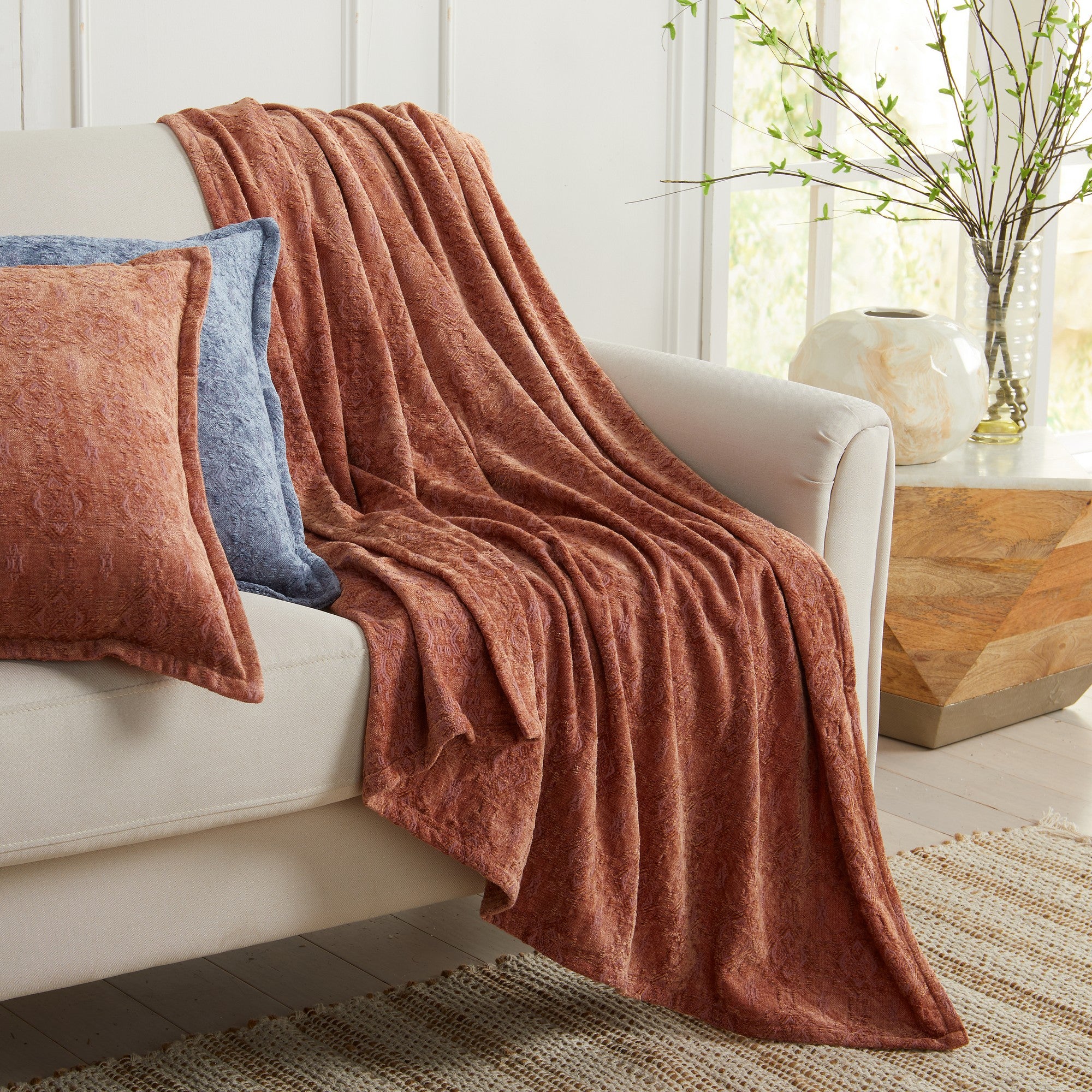 Clay Charlotte Collection chenille jacquard throw blanket draped over a cream sofa with coordinating clay and blue accent pillows in a bright living room setting.