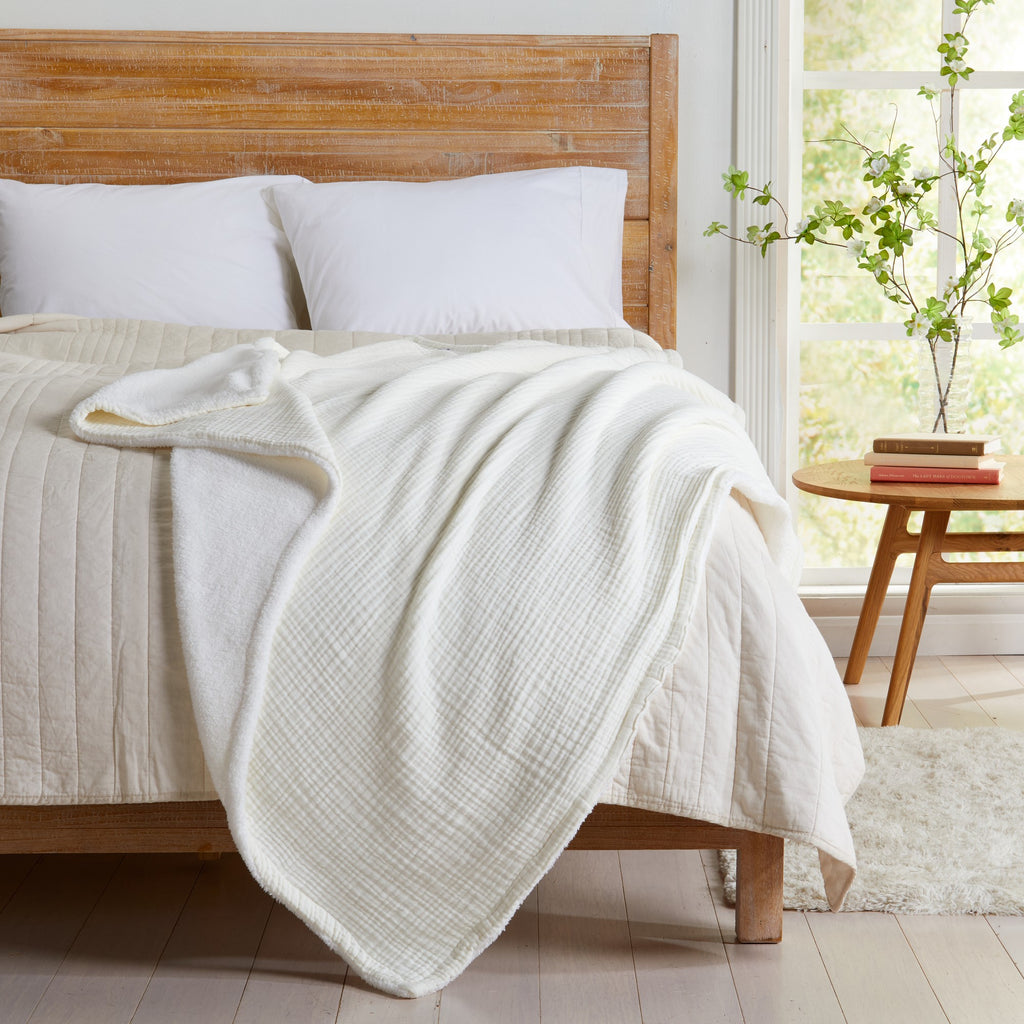 Off-white Maria Collection cotton muslin sherpa throw blanket draped across a bed, showcasing the textured cotton muslin face and soft sherpa backing in a bright bedroom setting.