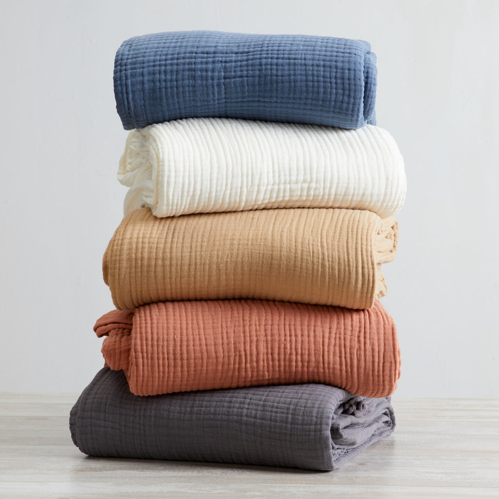 A stack of folded cotton muslin sherpa throw blankets in various solid colors including white, blue, beige, rust, charcoal, and orange.