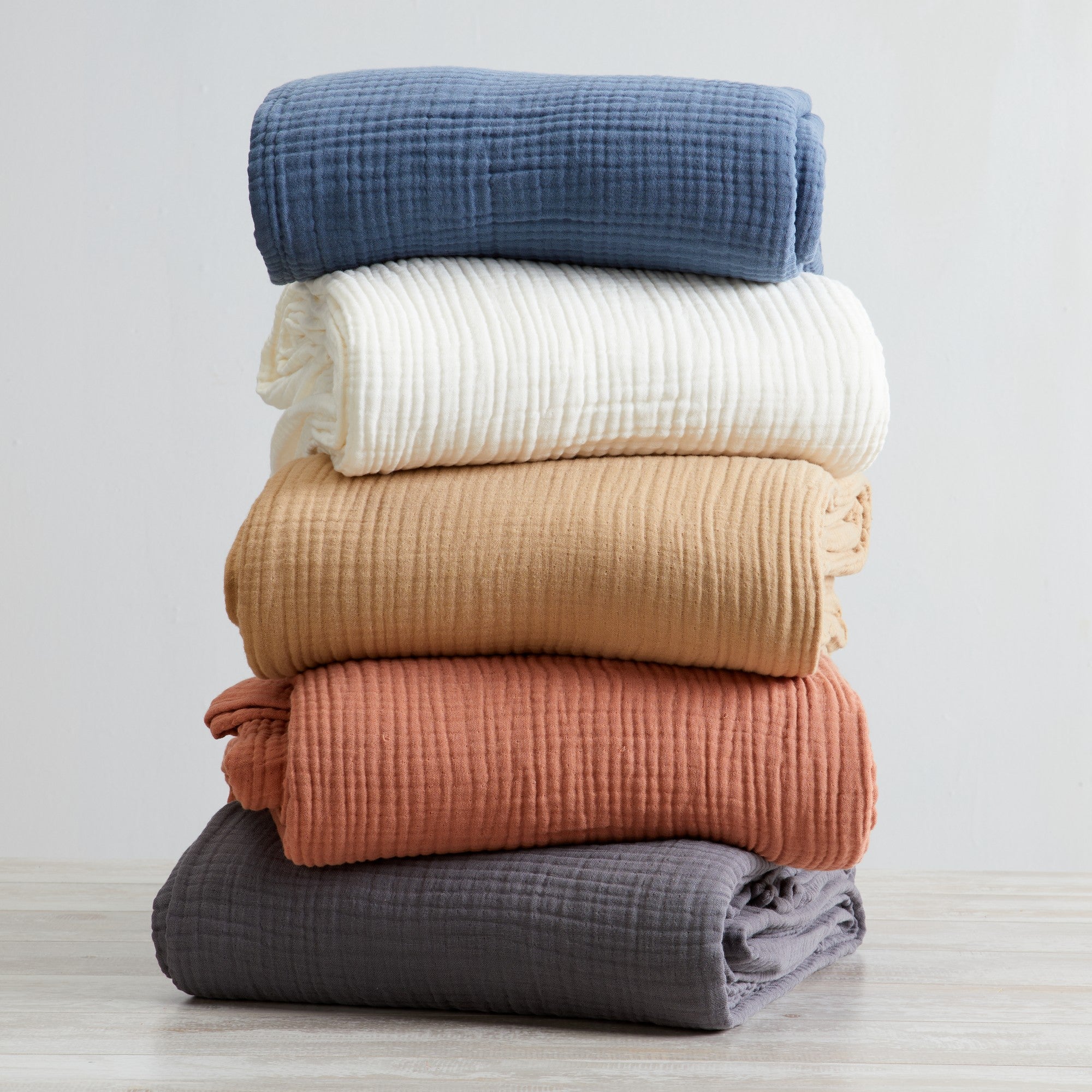 A stack of folded cotton muslin sherpa throw blankets in various solid colors including white, blue, beige, rust, charcoal, and orange.