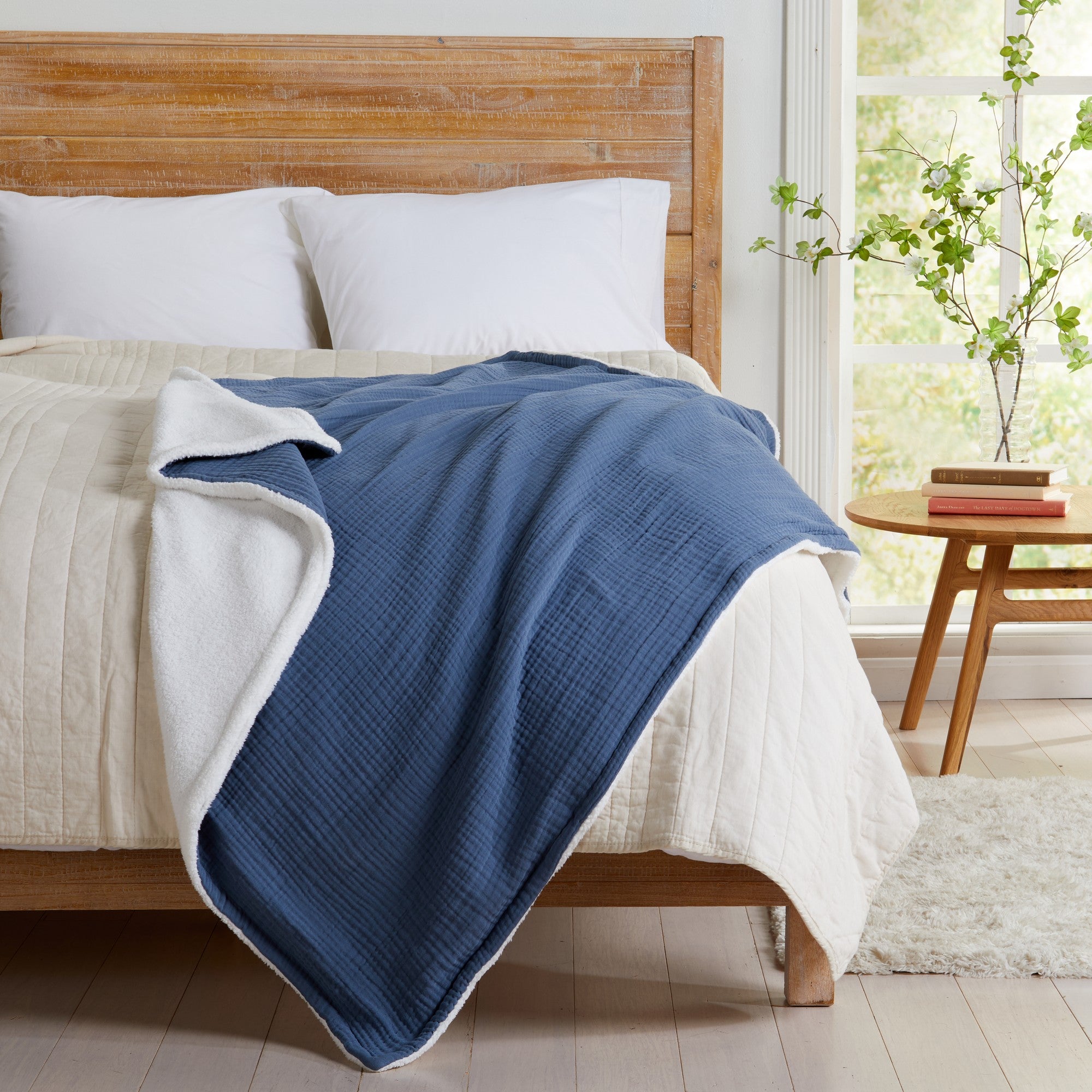 Blue Maria Collection cotton muslin sherpa throw blanket draped across a bed, showcasing the textured cotton muslin face and plush sherpa backing in a bright bedroom setting.
